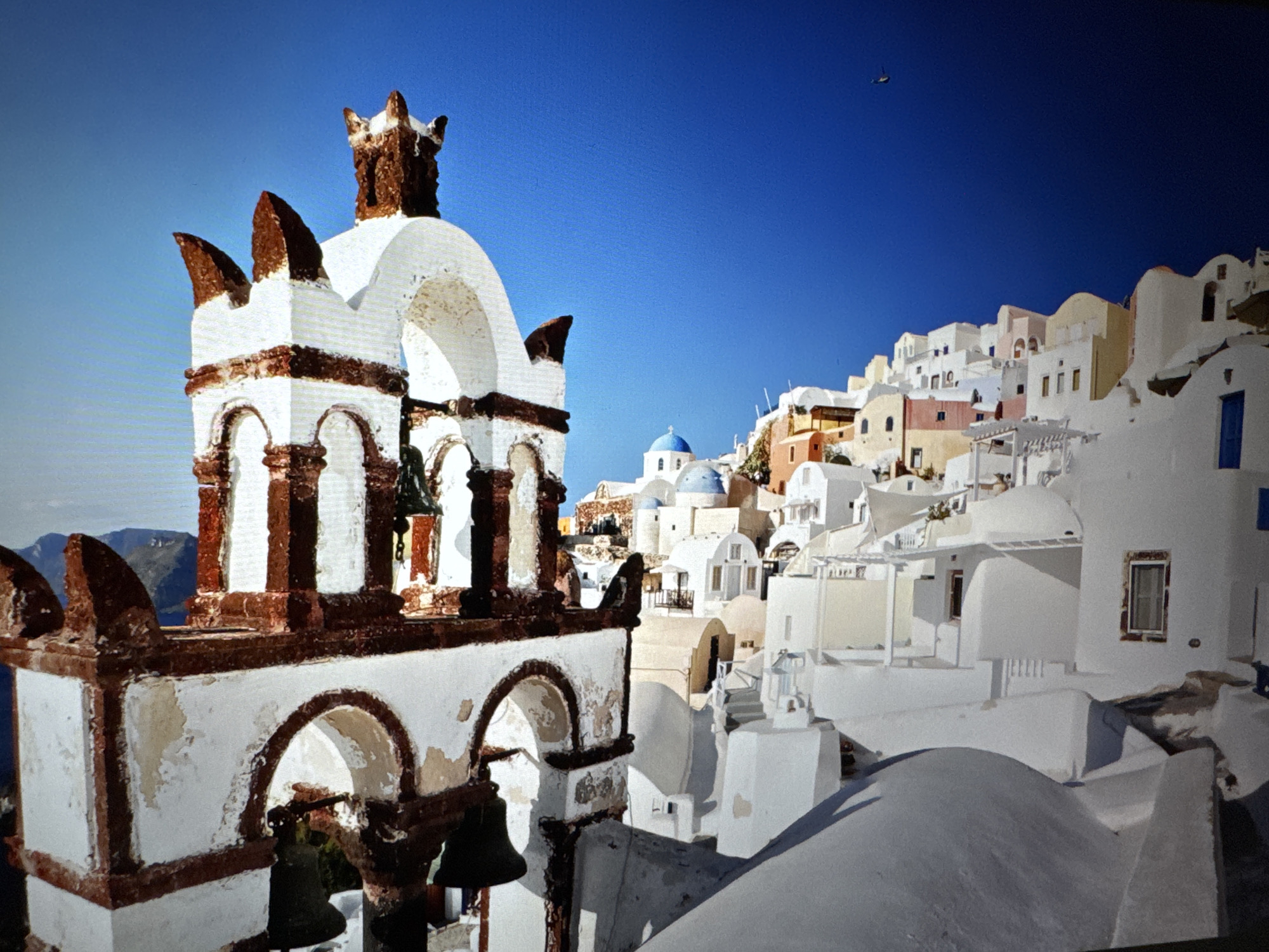 This vibrant image captures a picturesque scene of an iconic Greek island. In the foreground, prominently positioned on the left, is a traditional white church bell tower with distinctive dark reddish-brown decorative trim and arches. Several dark bells hang within its open structure.
Beyond the bell tower, a cascade of white Cycladic buildings with flat roofs and some with traditional blue domes stretch across the hillside to the right. The buildings vary in height and orientation, creating a tiered effect. Interspersed among the predominantly white structures are a few buildings with pastel-colored facades, such as light yellow, pale pink, and earthy brown tones. Blue doors and window frames add splashes of color.
The background features a strikingly clear, deep blue sky, indicating bright, sunny weather. A small, dark helicopter is visible high in the sky towards the upper right corner. In the far distance on the left, beyond the bell tower, faint outlines of mountains or hills can be seen under the bright sky. The overall impression is one of a serene and classic Mediterranean landscape. The time of day appears to be bright daylight, likely mid-morning or early afternoon, with distinct shadows suggesting a high sun. No visible text is present in the image. The architectural style strongly suggests the location is Santorini, Greece.