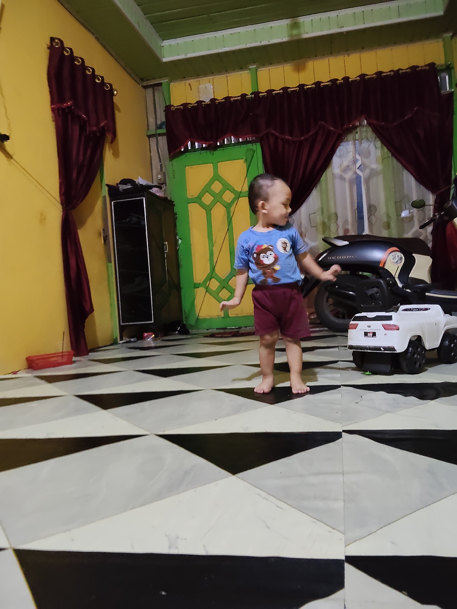 A young child is standing on a black and white tiled floor in an indoor room. The child is wearing a blue t-shirt with a monkey graphic and maroon shorts. To the child's right is a white toy car. Behind the child and the toy car is a dark-colored motorcycle. The room has yellow walls and green trim. There are dark red curtains hanging from the ceiling on the left and center of the image, and a green door with a patterned design is visible in the background. A dark wardrobe with reflective doors is located to the left of the child. The scene appears to be a domestic environment.