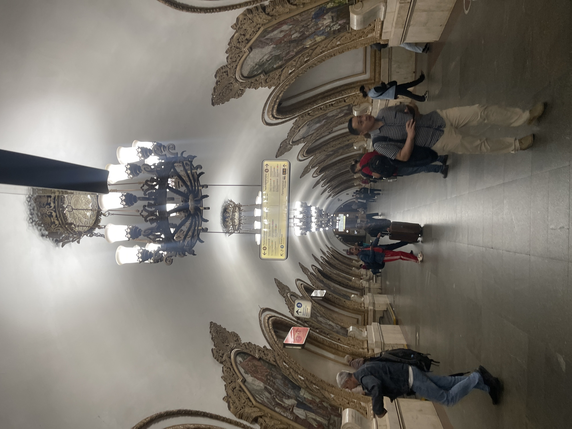 This image depicts the ornate interior of a Moscow Metro station, likely a historic one given the architectural style. The station features a high, vaulted ceiling illuminated by large, elaborate chandeliers. Along the sides of the tunnel, decorative arches frame large mosaic panels, which are richly ornamented with golden detailing. Several individuals are visible walking along the station platform. One man in the foreground is wearing a striped shirt and tan pants, walking towards the viewer. Other people, some carrying luggage, are scattered throughout the scene, indicating movement and transit. Signage in Cyrillic script is visible on the walls, providing directional information for passengers. The overall impression is one of grandeur and historical significance, characteristic of many of Moscow's older metro stations.