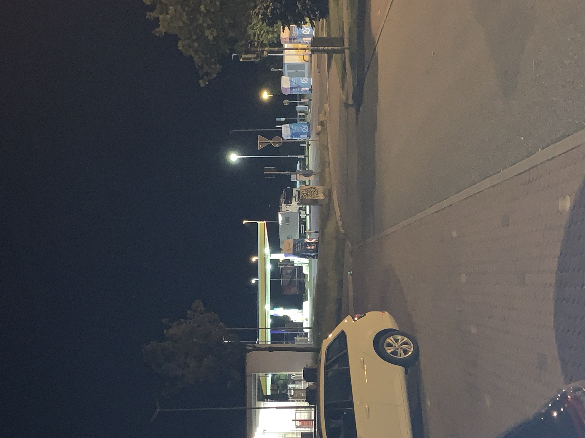 The image depicts a dimly lit outdoor scene at night, likely a rest stop or service area. A white station wagon is parked in the foreground, facing away from the camera. In the background, a gas station with illuminated pumps and a canopy is visible. Several large blue and white structures, possibly portable restrooms or waste disposal units, are also present in the midground. A portion of a truck is visible to the right of the gas station. The environment appears to be paved with concrete and asphalt, with some grassy areas and trees. The sky is dark and clear. The overall impression is that of a functional, utilitarian area, possibly along a highway or in a commercial zone, in Siemz-Niendorf, Germany.