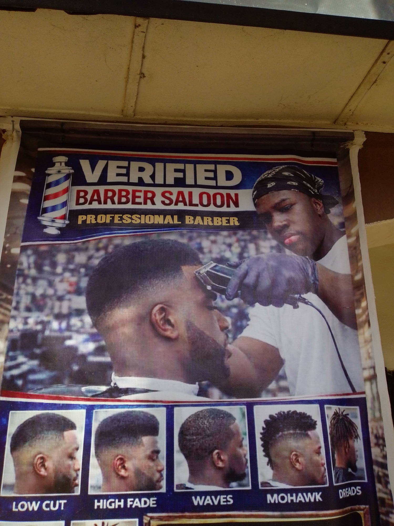 This is a photograph of a barber shop advertisement poster, likely located in Jalingo, Nigeria, as indicated by a previous query. The poster features a barber, wearing a bandana and gloves, giving a haircut to a client, with a barber pole graphic on the left. Below the main image, there are smaller photos showcasing various hairstyles like "LOW CUT," "HIGH FADE," "WAVES," "MOHAWK," and "DREADS." The text on the poster reads "VERIFIED BARBER SALOON PROFESSIONAL BARBER." The poster is attached to a structure, possibly a building or a stall, with a textured ceiling visible at the top. The lighting appears to be natural daylight.