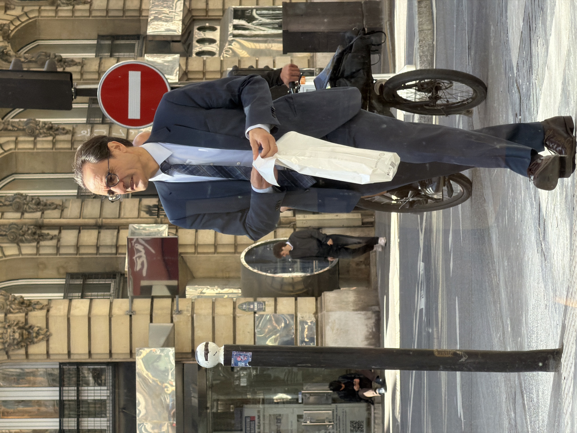 A man in a navy suit stands next to a vintage-style motorcycle on a street in Paris, France. He holds a white bag in his left hand, and is looking to his left. The background features a building with ornate architectural details and a traffic sign. A pole is on the right side of the image. The street has visible lane markings. Other people are present in the background.