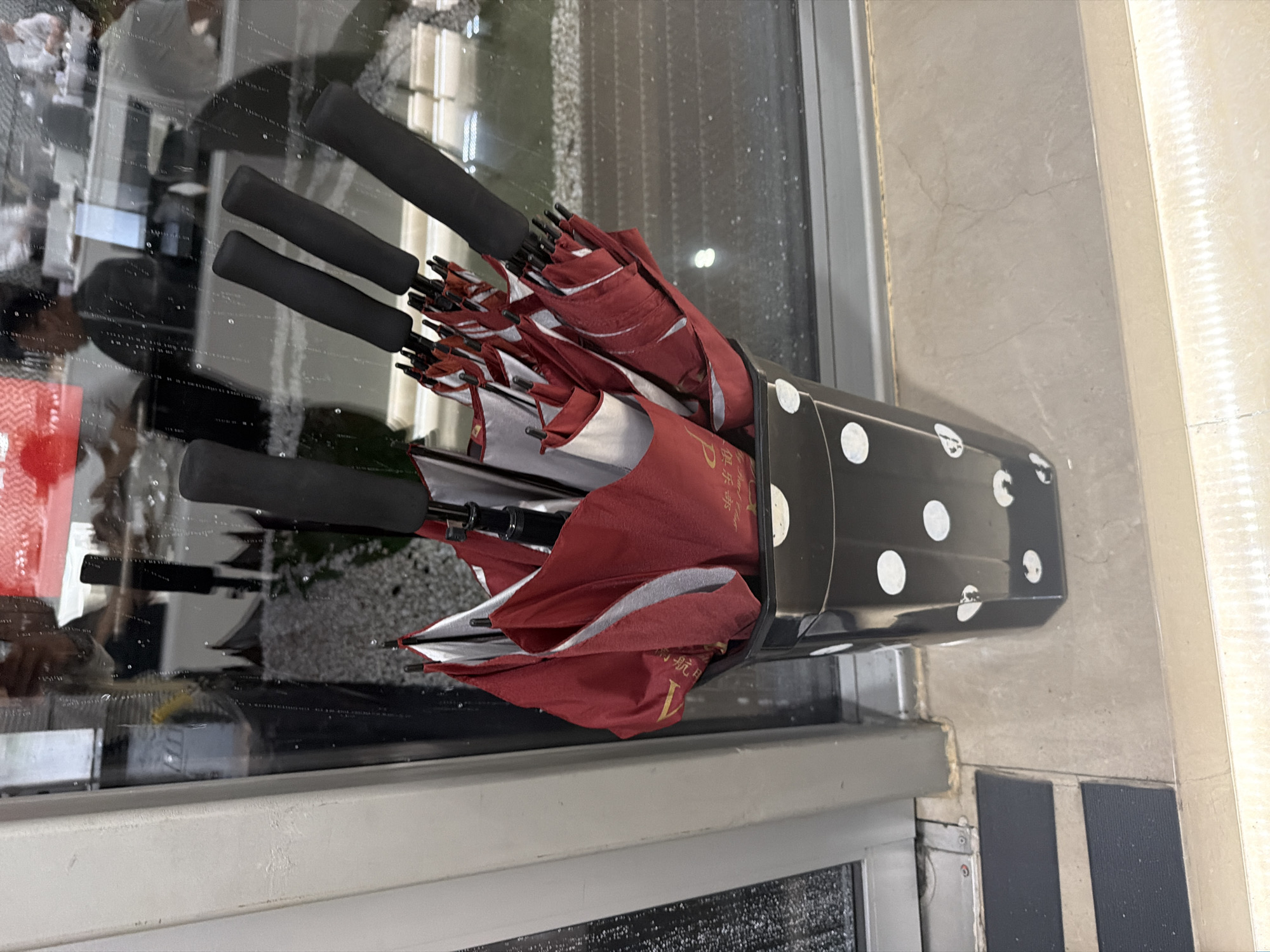 An umbrella stand filled with numerous red and white folded umbrellas is positioned next to a large glass surface and a light-colored tiled floor or wall. The image is taken from a slightly elevated perspective, looking down.
The umbrella stand is black with white polka dots of varying sizes. Inside, several closed umbrellas are neatly stored, with their black handles pointing upwards. The umbrellas are primarily red on the exterior, with white or silver lining visible on the inside of the folded fabric. Some of the red fabric features golden text, which appears to be Chinese characters. Visible characters include "航" (háng), "天" (tiān), "品" (pǐn), and an English letter that looks like "L" or "Y," suggesting a brand name or promotional message.
The glass surface, likely a window or door, reflects the interior space, showing blurry shapes of people and objects, and possibly some distant outdoor elements. There are numerous small smudges or droplets on the glass. The light-colored tiled floor or wall on the right side of the frame exhibits a subtle pattern of cracks or grout lines. Near the bottom edge, dark, textured strips are visible on the ground, possibly non-slip mats. The presence of multiple umbrellas indicates a setting where protection from rain is provided or anticipated, possibly an entrance to a public building such as a hotel, office, or shopping center. The lighting suggests it is daytime.