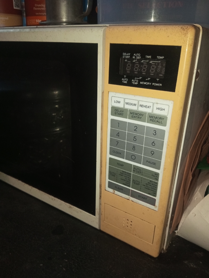 An old, yellowed microwave oven is shown, partially obscured by objects on the side. The control panel features a digital display showing "88:88", buttons for power levels (LOW, MEDIUM, REHEAT, HIGH), numbered keypad, and function buttons like DELAY START, MEMORY ENTRY, MEMORY RECALL, CLOCK, PAUSE, TEMP, AUTO W-DEF, AUTO TEMP, CLEAR, and START. Labels detail automatic cooking functions like bacon, baked potatoes, canned vegetables, and frozen dinners. The oven's exterior exhibits signs of wear and age.