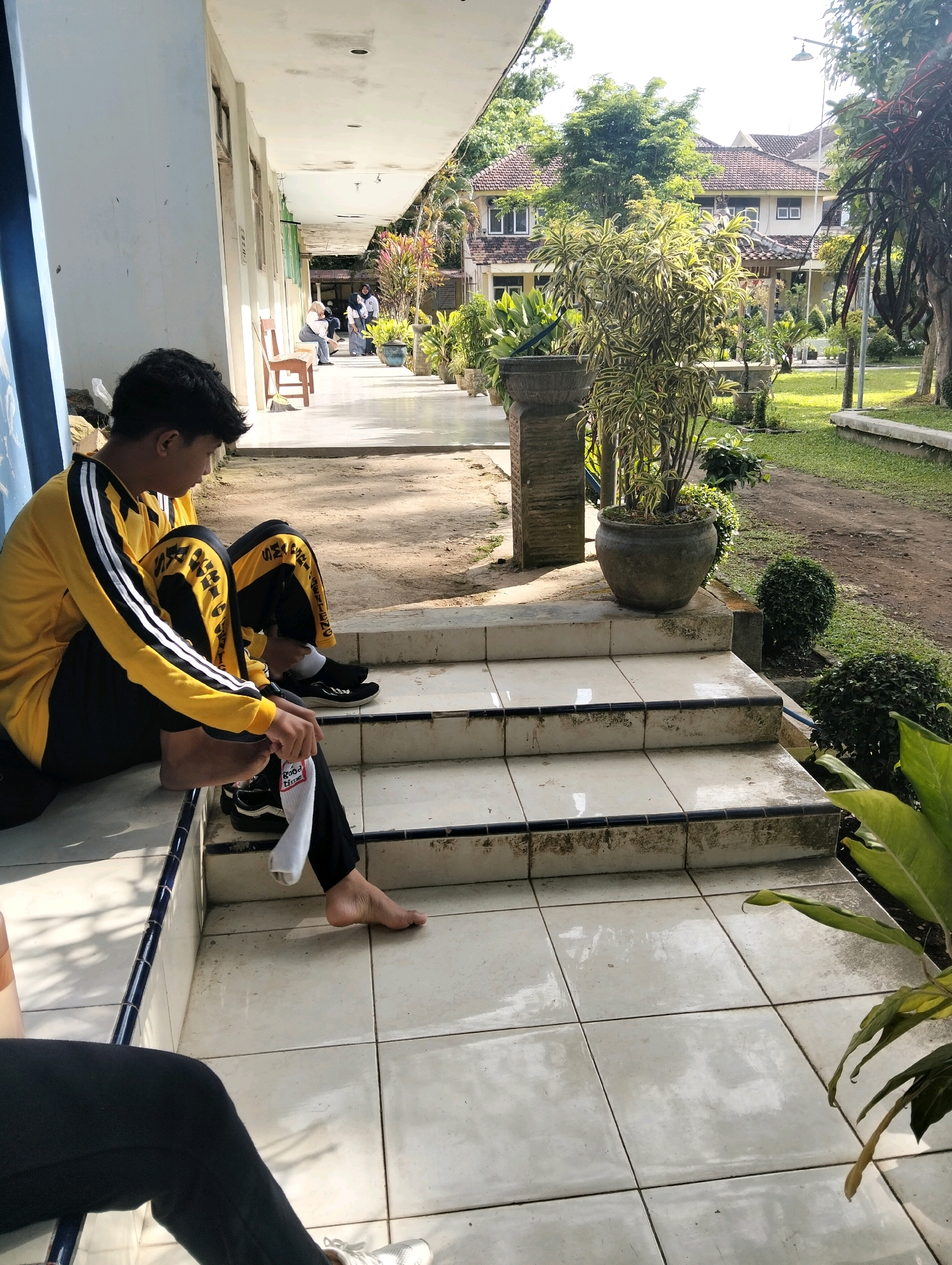 A young person, appearing to be male, is sitting on the ground next to a set of tiled stairs. They are wearing a yellow long-sleeved shirt with black and white stripes on the sleeves and black pants with yellow accents. Their right leg is extended forward, and their left leg is bent with their foot resting on the ground. A white sock is pulled down on their right foot, and a black sneaker is visible on their left foot. The individual is looking downwards. The environment appears to be an outdoor walkway or patio area with a tiled floor and steps. In the background, there is a building with a red tiled roof, trees, and lush green vegetation. The scene is illuminated by natural daylight. The location is identified as Genteng, Indonesia.