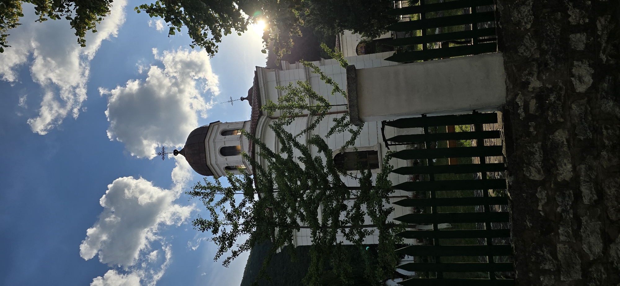 An exterior, eye-level, vertical shot captures a white church with a copper dome against a blue sky dotted with fluffy white clouds. Sunlight beams from the right side, partially obscuring the view with lens flare. Green trees with dark leaves frame the scene on the left and right. A dark green, pointed-top wooden fence runs across the foreground, with a stone wall beneath it. A white pillar stands in front of the fence, slightly to the right of the center. The church features arched windows and multiple crosses on its roof. The background shows a lush, green hillside.