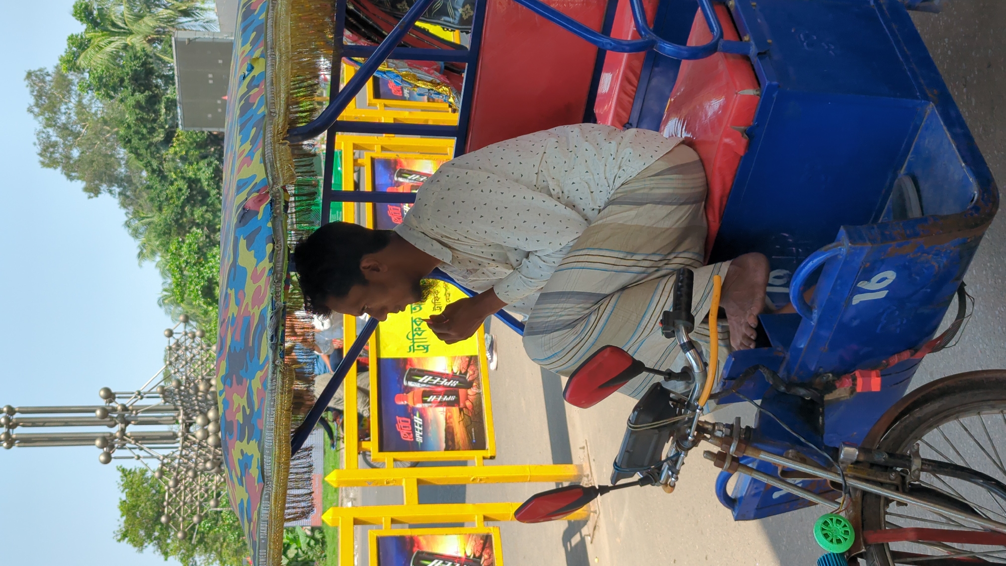A barefoot, seated man in a white patterned shirt and striped lungi is in a blue and red rickshaw with yellow framing near the Shahid Minar monument in Dhaka, Bangladesh. He is looking down and appears to be eating. Advertisements for "SPEED" energy drinks are visible on the rickshaw’s frame. The rickshaw has side mirrors and the number "16" on its side. Vegetation and a light blue sky are in the background.