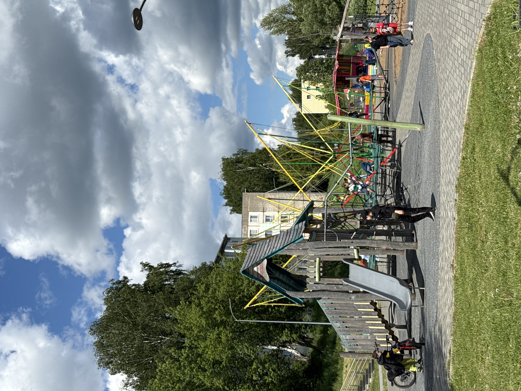The image shows a playground in Votkinsk, Russia. The playground has a slide, a climbing structure made of ropes, and a merry-go-round. People are using the playground equipment. There are trees and buildings surrounding the playground. A cloudy sky is visible in the background. A parked bicycle is near the slide.