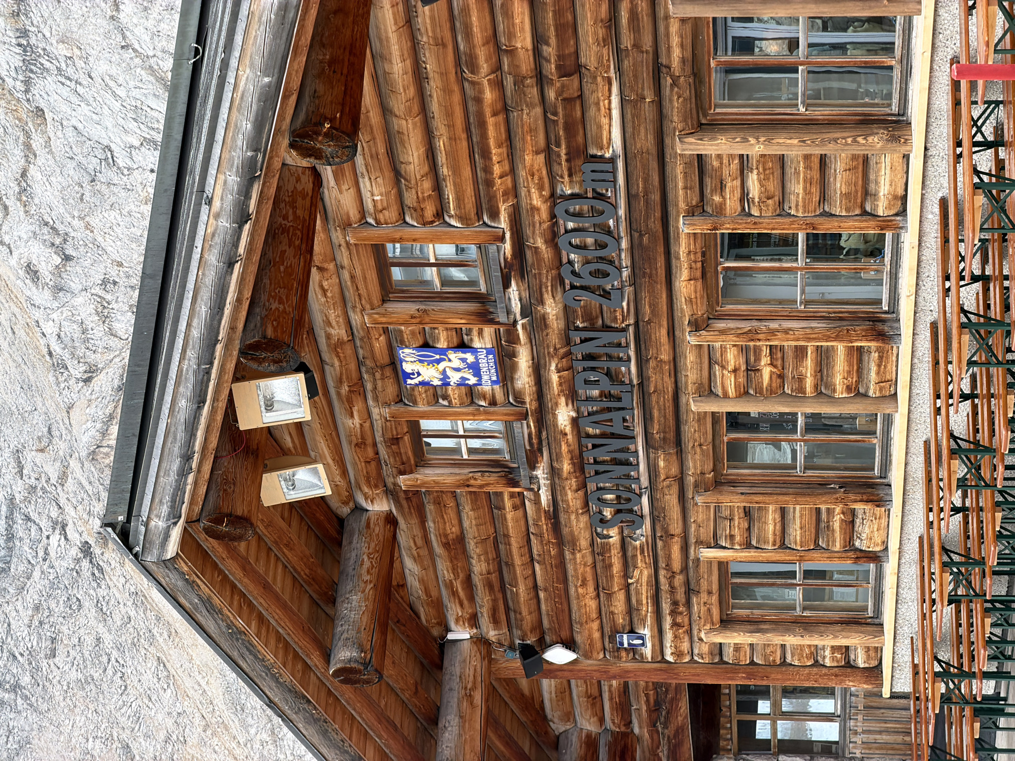 The image shows a rustic, wooden building with "SONNALPIN 2600m" written in black lettering on its facade. The building appears to be a mountain hut or restaurant, possibly located at a high altitude given the "2600m" inscription, which could indicate an elevation of 2600 meters above sea level. Behind the building, a rugged, rocky mountain slope is visible, suggesting a mountainous environment. The building has several windows and two outdoor lights mounted on the upper section. In front of the building, there are rows of empty wooden picnic tables with green metal legs, indicating an outdoor seating area. A blue and yellow crest with a lion and text reading "LOWENBRAU MUNCHEN" is mounted on the wall above one of the windows. This crest suggests a connection to the Löwenbräu brewery from Munich. The overall scene conveys a sense of being in a mountainous region, likely a tourist destination for outdoor activities. While the specific location within Garmisch-Partenkirchen, Germany is not explicitly stated, the presence of "MUNCHEN" on the crest and the mountainous landscape are consistent with an Alpine setting in or near the Bavarian Alps, which includes Garmisch-Partenkirchen.