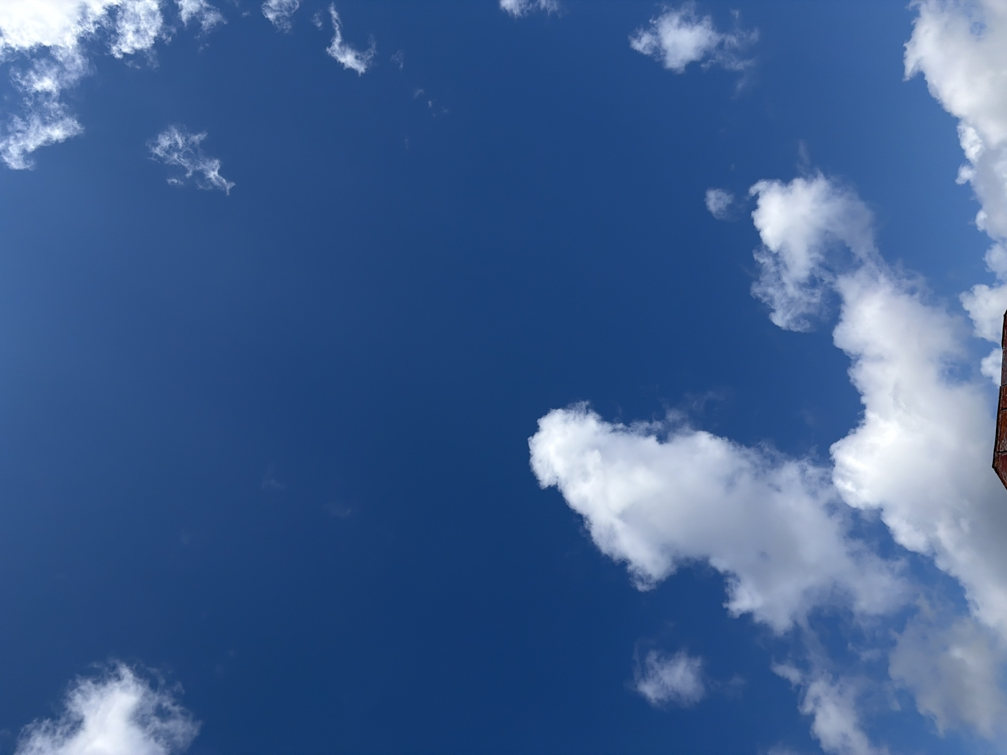 The image captures a clear blue sky with scattered white cumulus clouds. In the lower portion of the frame, a dark red brick rooftop is partially visible, suggesting the presence of a building. The scene appears to be an outdoor environment, likely within a city, given the rooftop. There are no people, animals, or visible actions depicted in the image. The overall impression is one of a bright, sunny day. Without additional information or context, it is not possible to determine the specific location within the city of Chernihiv, Ukraine.