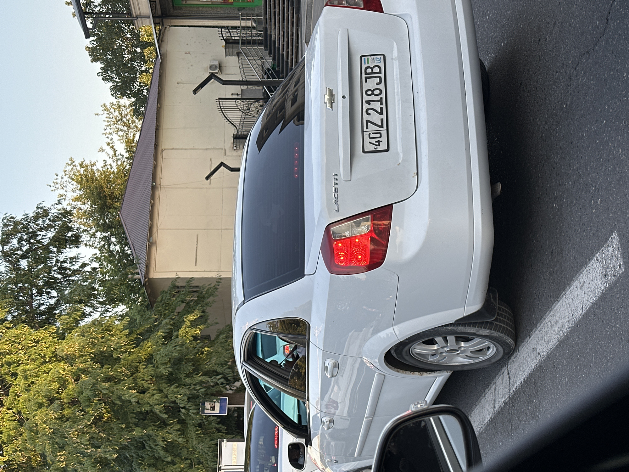 A white Chevrolet Lacetti is parked on a city street in Tashkent, Uzbekistan. The car has tinted windows and a license plate that reads "40 Z 218 JB". In the background, there are trees, buildings, and a pedestrian walkway with stairs. The scene appears to be during the day.