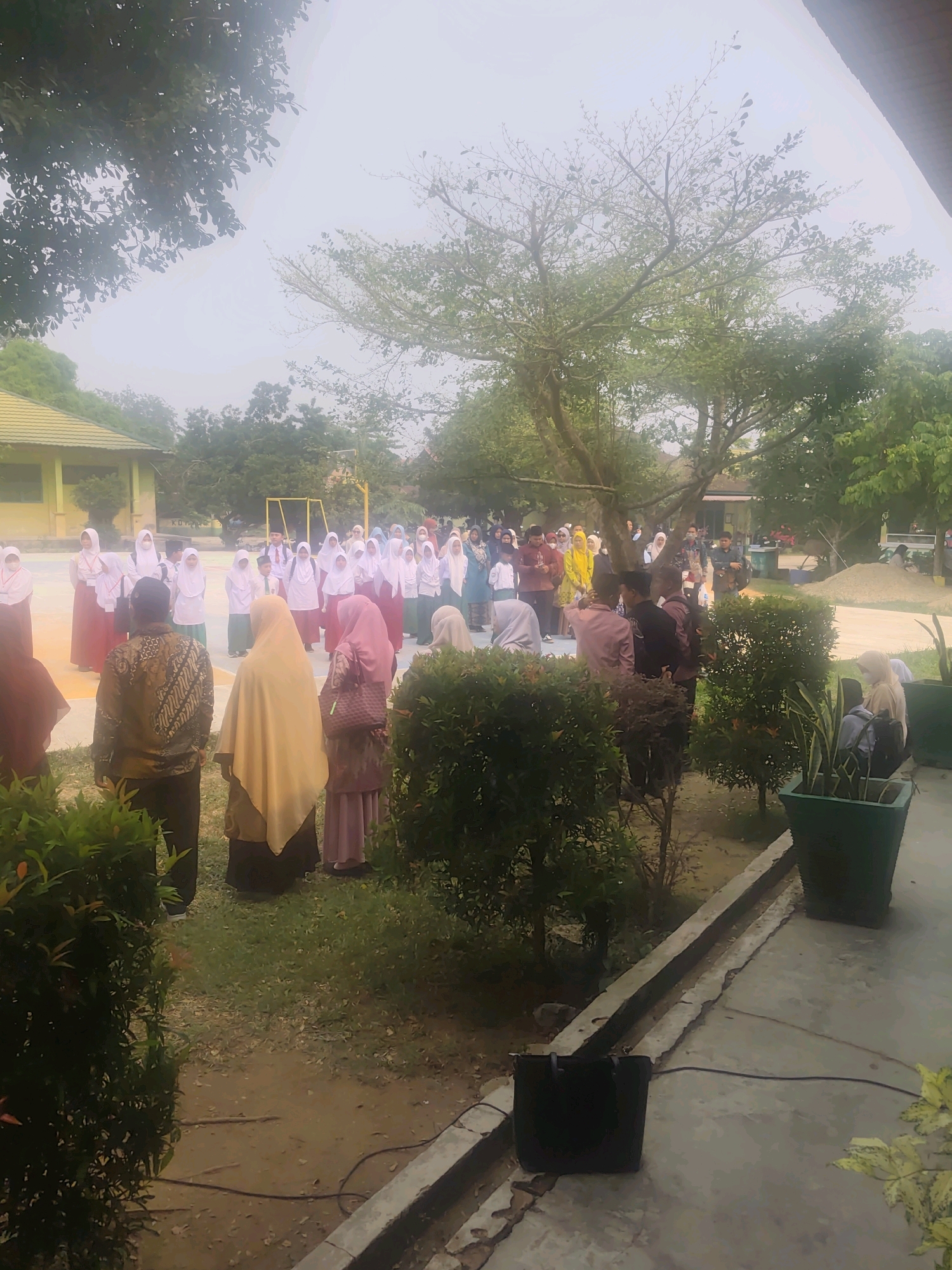 The image shows an outdoor gathering of people, likely students and adults, in what appears to be a schoolyard in Pematang Reba, Indonesia. In the foreground, three adults are seen from behind, facing a group of predominantly female students. The students, many wearing hijabs and uniforms, are gathered in the background, appearing to be in an assembly or a formal event. One adult in the foreground wears a batik-patterned shirt. Behind the students, a building with a yellow roof is partially visible, and trees are scattered throughout the scene. The ground is a mixture of paved areas and dirt/grass. The lighting suggests it is daytime, possibly overcast. There are also several potted plants in the foreground and midground, and a black bag is placed on the ground near the paved walkway. The overall atmosphere seems orderly and communal.