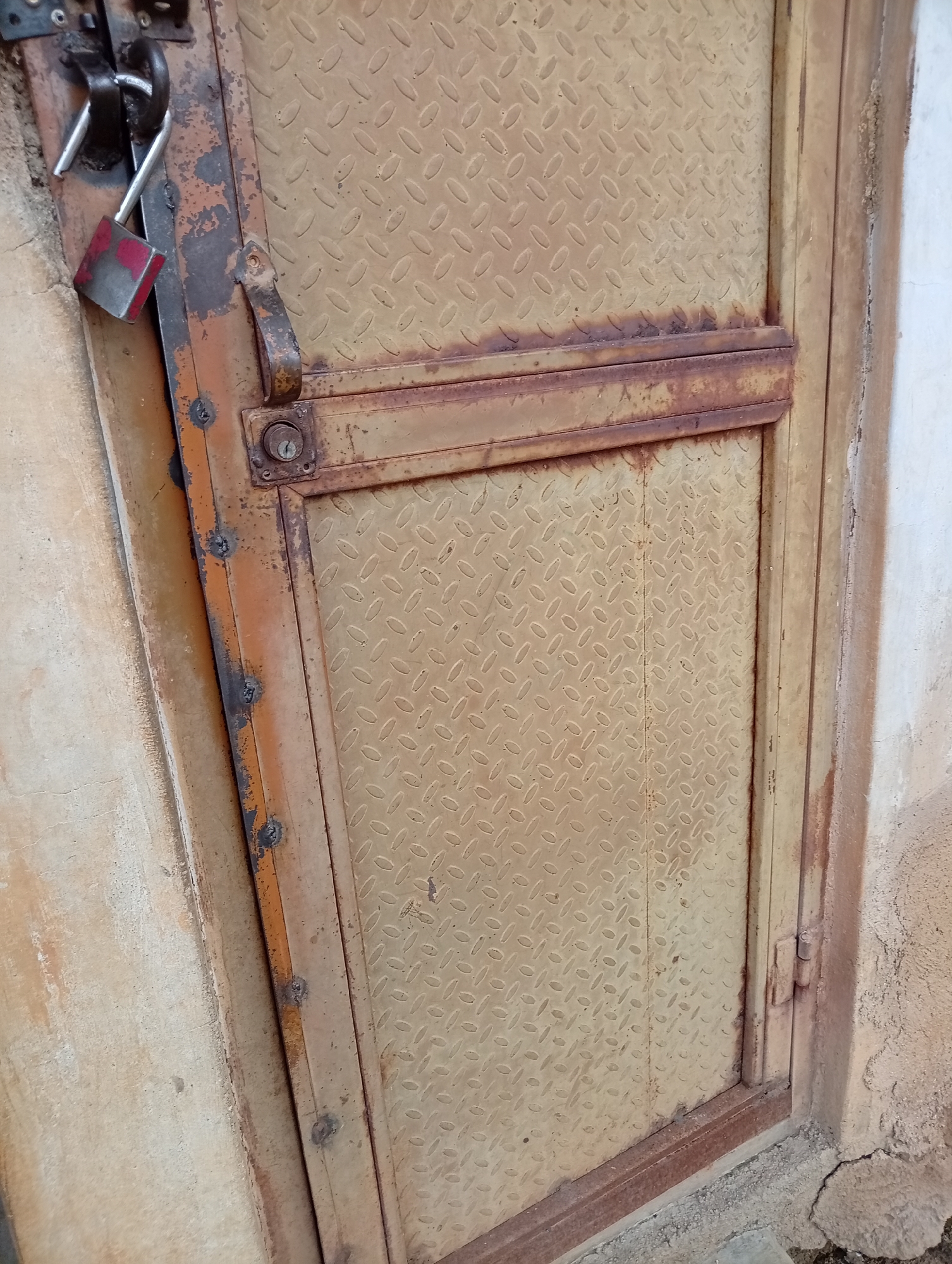 The image shows a close-up of a rusted metal door with a diamond plate pattern. A red and silver padlock is attached to a metal loop on the left side of the door frame, with the shackle in the open position. The door appears to be old and weathered, with peeling paint and visible signs of rust. The surrounding wall is a rough, textured, light-colored material. The scene is likely outdoors, given the apparent weathering of the door and the rough texture of the wall, possibly an entrance to a building or an enclosure. The image is taken from a slightly downward angle, focusing on the lock and the upper portion of the door.