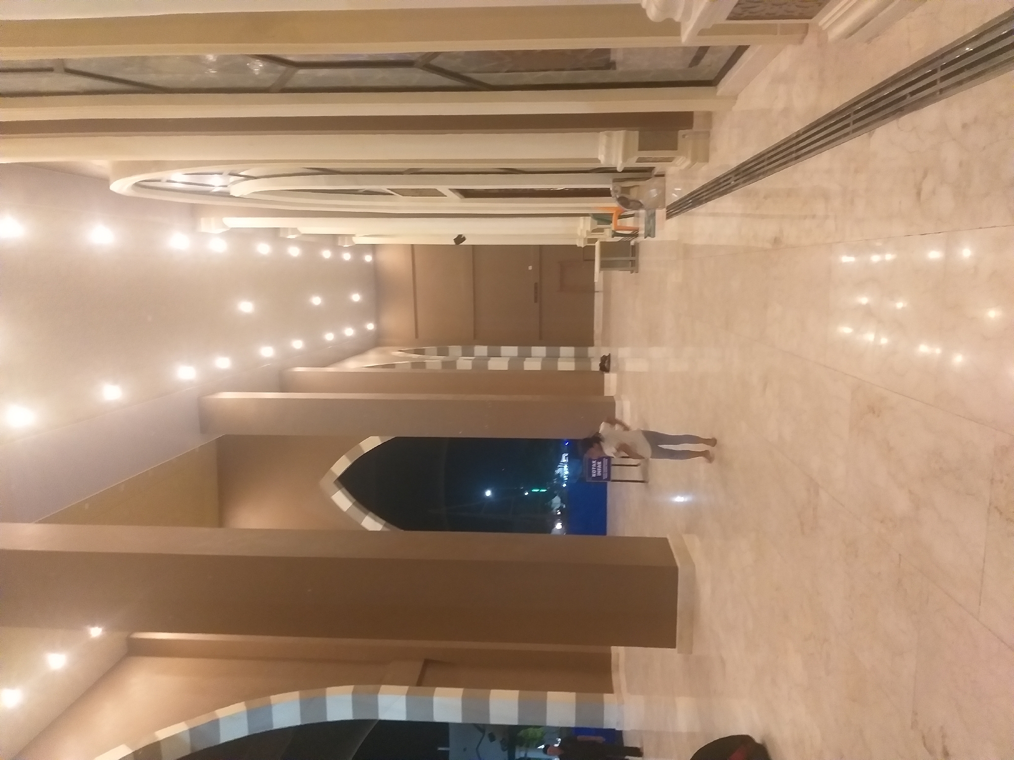 The image shows a wide, marble-floored corridor in a building that appears to be a mosque or similar public building in Dumai City, Indonesia. The architecture features large, brown pillars and arched doorways with white detailing. The ceiling is lit by numerous recessed lights. A young girl in a white shirt and grey leggings stands in the center of the corridor, facing away from the camera. To the left, a man in dark clothing walks away from the camera. A sign with the text "KOTAK INFAR" is visible near the girl. The environment is clean and well-maintained, with reflections on the polished floor.