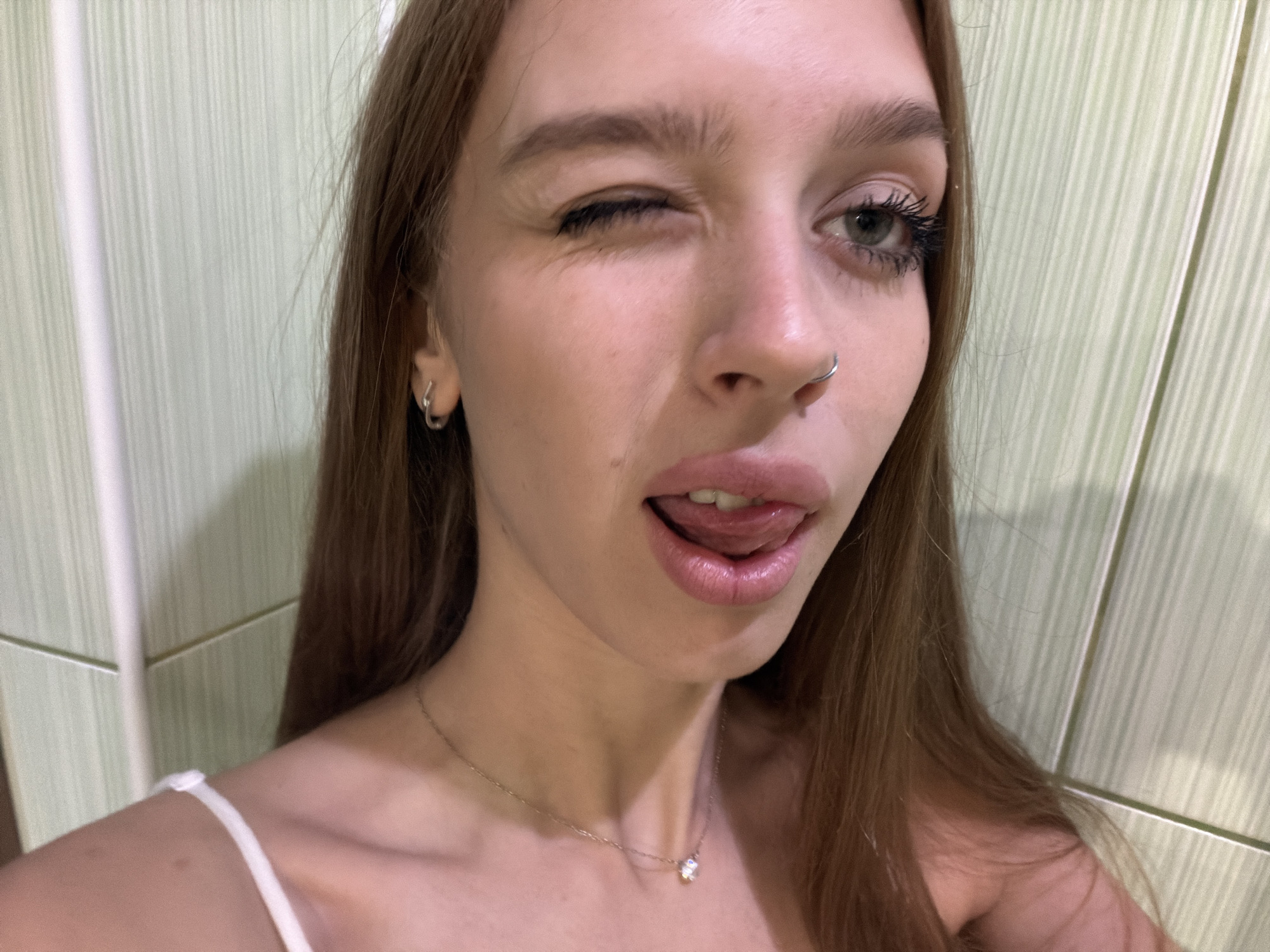 The image is a close-up selfie of a young woman. She has long, brown hair that falls around her shoulders and a fair complexion. She is wearing a white spaghetti strap top and a delicate necklace with a small pendant. She has a nose ring and a small hoop earring in her visible ear.
The woman is winking with her left eye, while her right eye is open and looking towards the camera. Her mouth is slightly open, and her tongue is peeking out, giving her a playful expression. The lighting is soft, and the background appears to be a wall with vertical green and white stripes, possibly in a bathroom or dressing area. The overall mood of the image is lighthearted and flirtatious, suggesting a moment of fun or silliness. There is no visible text in the image.