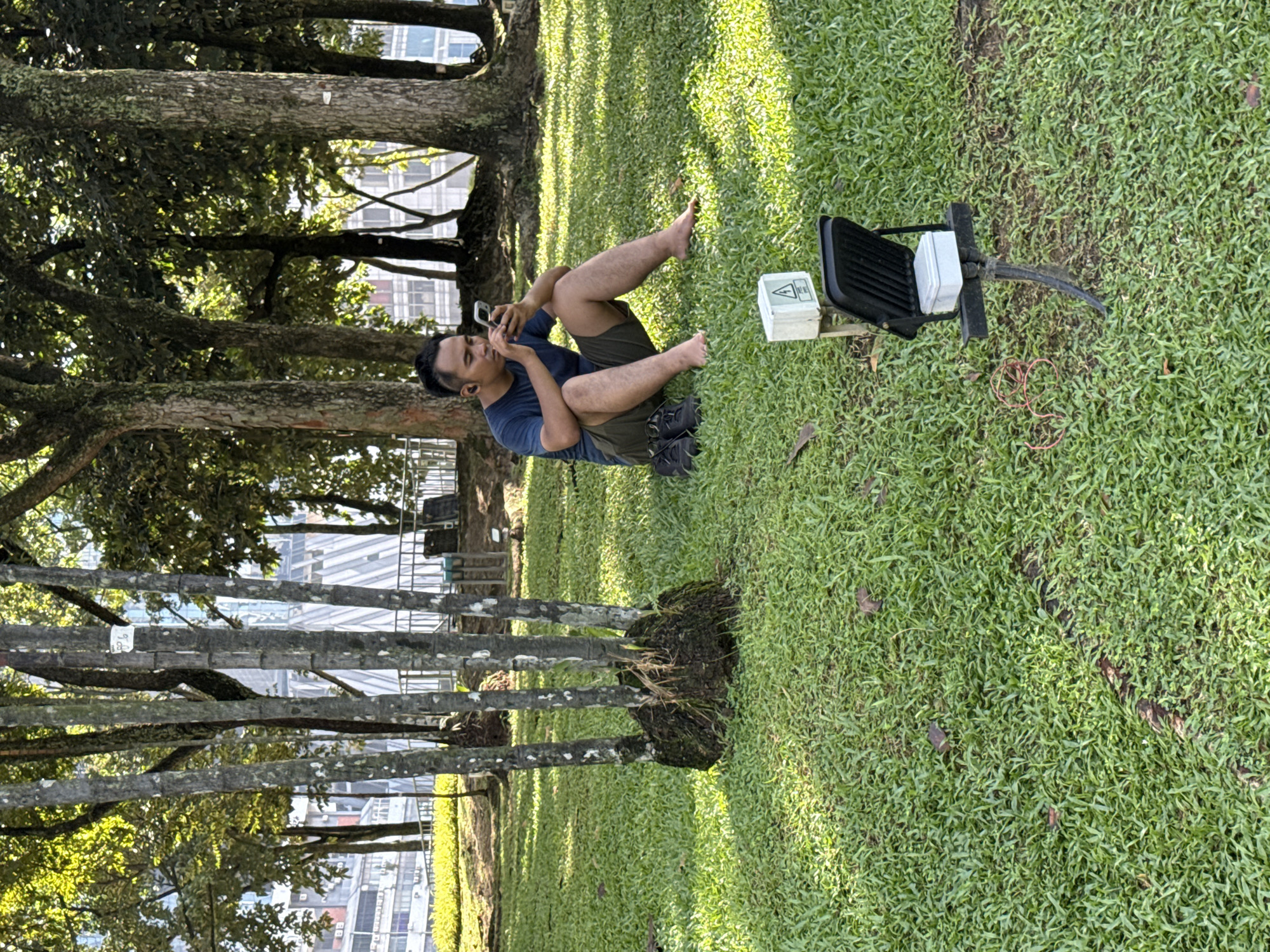 The image shows a man sitting on the grass in a park setting. He is barefoot, wearing a blue shirt and dark shorts, and looking at a smartphone. He appears to be in conversation. Beside him is a piece of equipment attached to an electrical junction box. In the background are trees and a tall building, suggesting an urban park in the city of Kuala Lumpur, Malaysia.