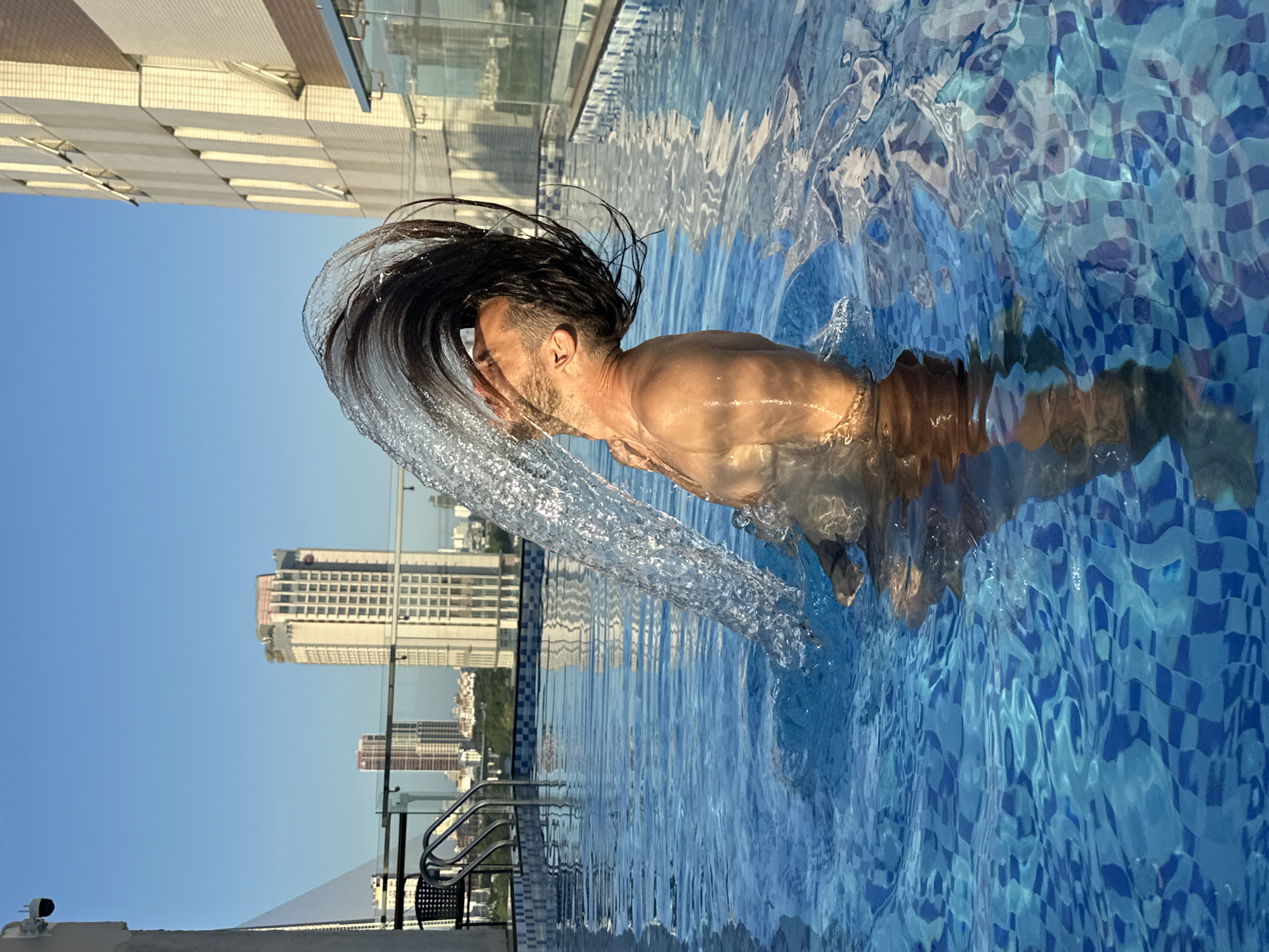 The image depicts a man in a swimming pool on a rooftop, with buildings and a clear blue sky in the background. The man, with dark hair, is captured mid-action, flipping his hair backwards, creating a splash of water that arcs upwards. His body is partially submerged in the pool, and sunlight illuminates his skin, highlighting his muscular physique. The pool itself has a tiled bottom with a blue and white checkered pattern. In the distance, several tall buildings are visible, characteristic of a city skyline. A railing and a ladder are present near the edge of the pool, suggesting it is part of a hotel or residential complex. The overall impression is one of leisure and dynamism, set against an urban backdrop. The presence of tall buildings and the general architectural style suggest a location in a city, and the hint provided in the prompt specifies Đà Nẵng, Vietnam.