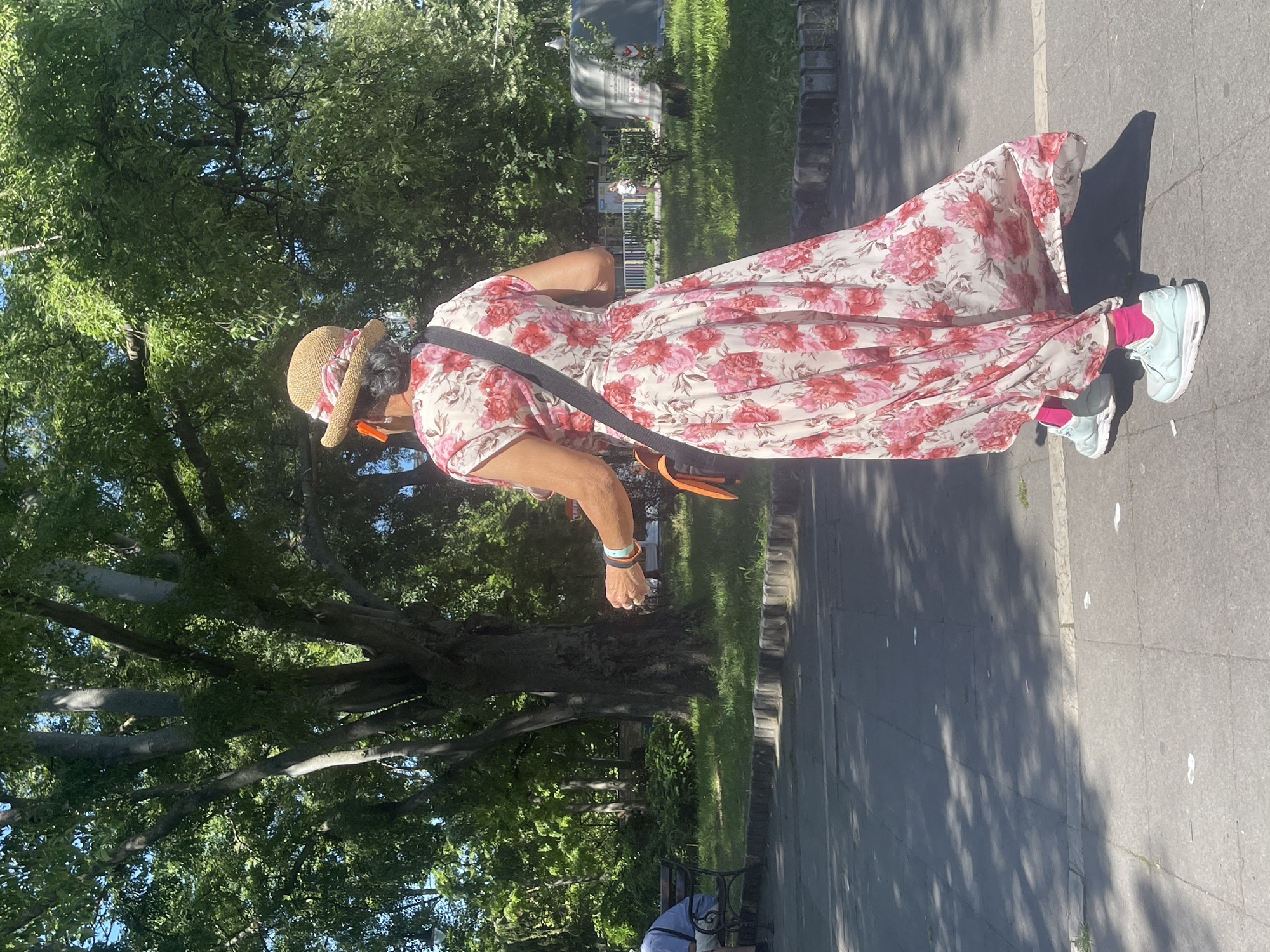 An older woman, viewed from behind, walks across a paved area in a park. She wears a straw hat with a patterned ribbon, a long, flowing dress with a floral print of pink and red flowers on a white background, and bright pink socks with light blue athletic shoes. She carries a black crossbody bag over her right shoulder and an orange purse or pouch hangs from her left side. She has a wristband on her left wrist. The park setting features a large, mature tree with lush green foliage, a lawn, and a paved path. In the background, a man sits on a park bench and other park elements are visible, including a large green trash receptacle. The image is taken on a sunny day, casting dappled shadows on the pavement. This scene is located in the Primorski Park in Varna, Bulgaria.