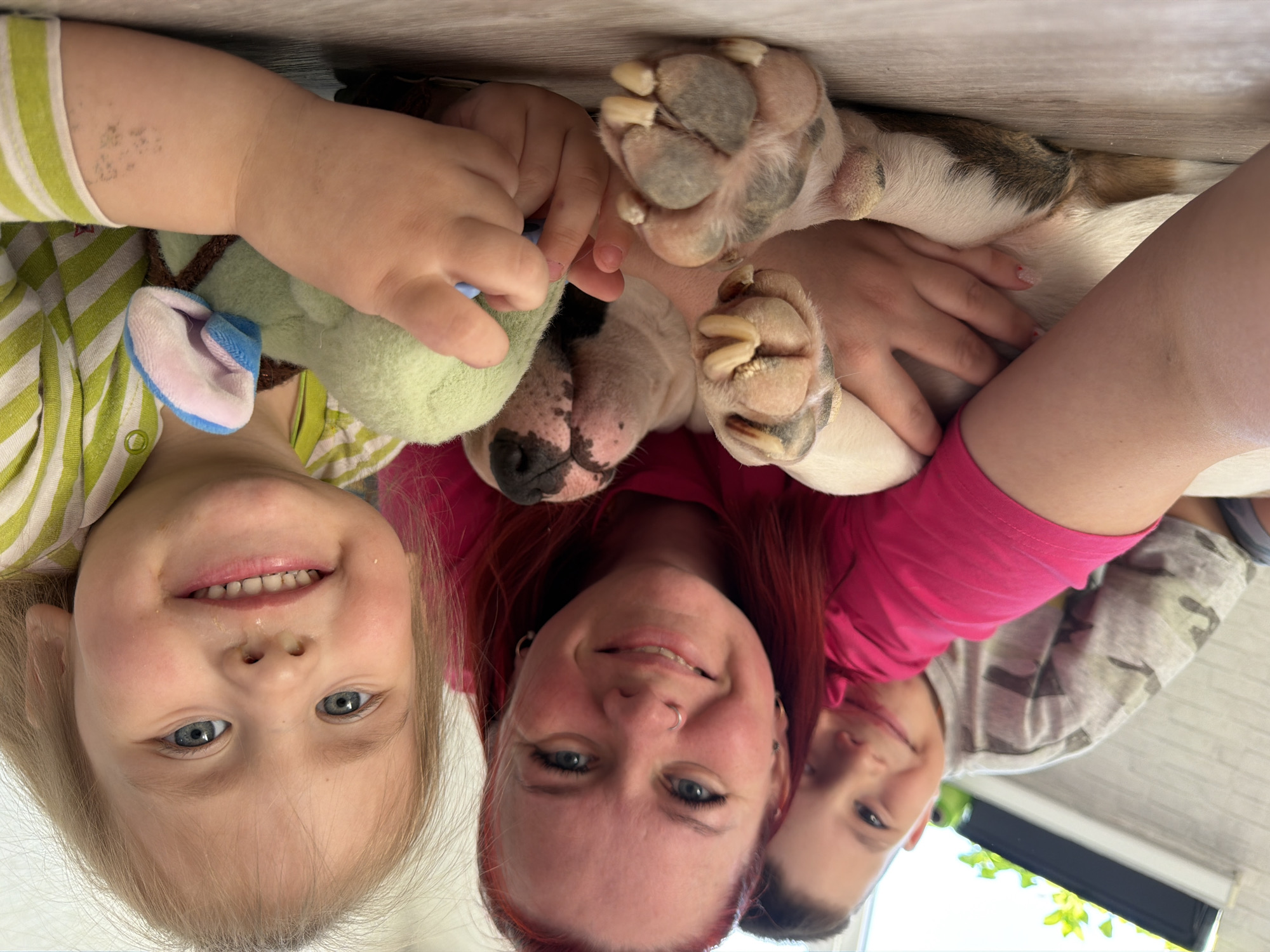 A photo, rotated 180 degrees, shows a close-up of a child, a woman, a boy, and a dog in an indoor setting. The child on the left wears a green and white striped shirt and holds a green stuffed animal. The woman in the center has red hair and a pink shirt. The boy on the right wears a camouflage shirt. The dog's paws are in the center of the frame. The background appears to be a wooden surface or floor. The location is not clear from the image and is not necessarily in Chaikovsky, Russia.