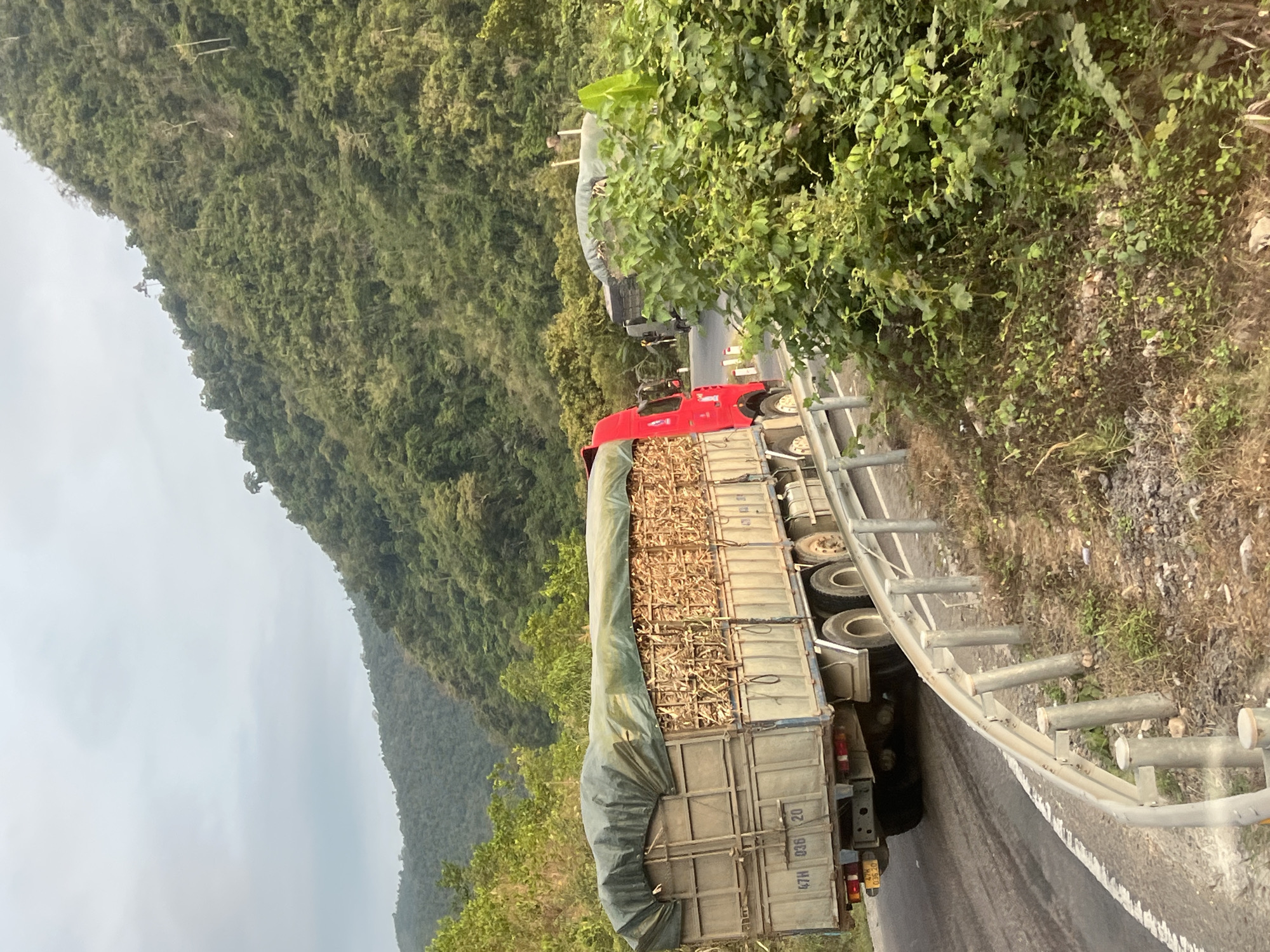 The image shows a truck on a winding road in Aê Thi, Vietnam. The truck is loaded with what appears to be a cargo of bamboo or similar material, partially covered by a tarp. The road is lined with a metal guardrail on one side. In the background, there is a steep, densely forested hillside. The weather seems overcast. Another truck is visible further up the road.