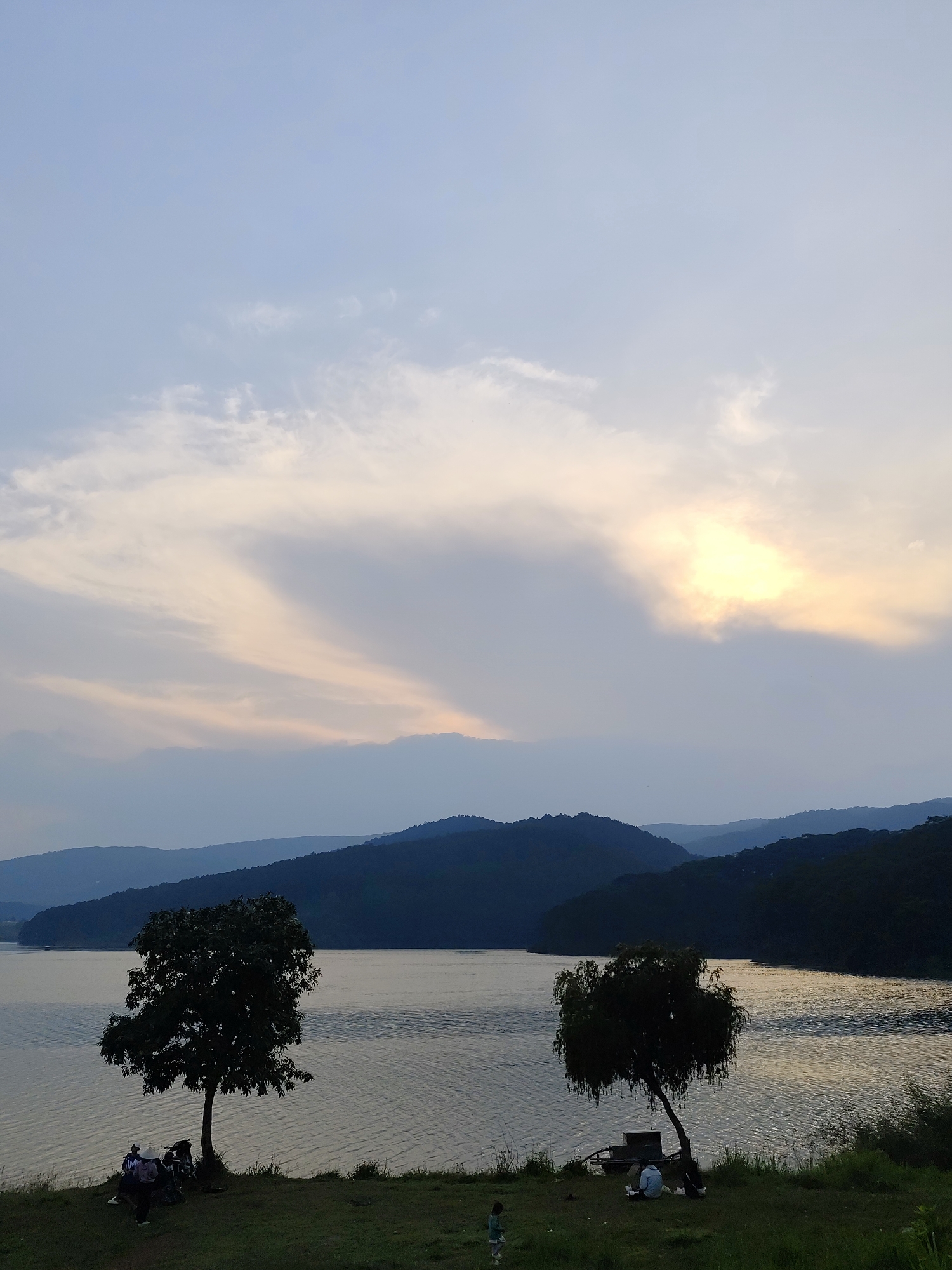 The image shows a scenic view of a lake surrounded by hills in Da Lat, Vietnam. The sky is overcast with a hint of sunlight breaking through the clouds. Two trees are in the foreground on a grassy bank, with small groups of people gathered near each tree. The lake's surface reflects the sky, creating a shimmering effect. The background features rolling hills covered in vegetation.