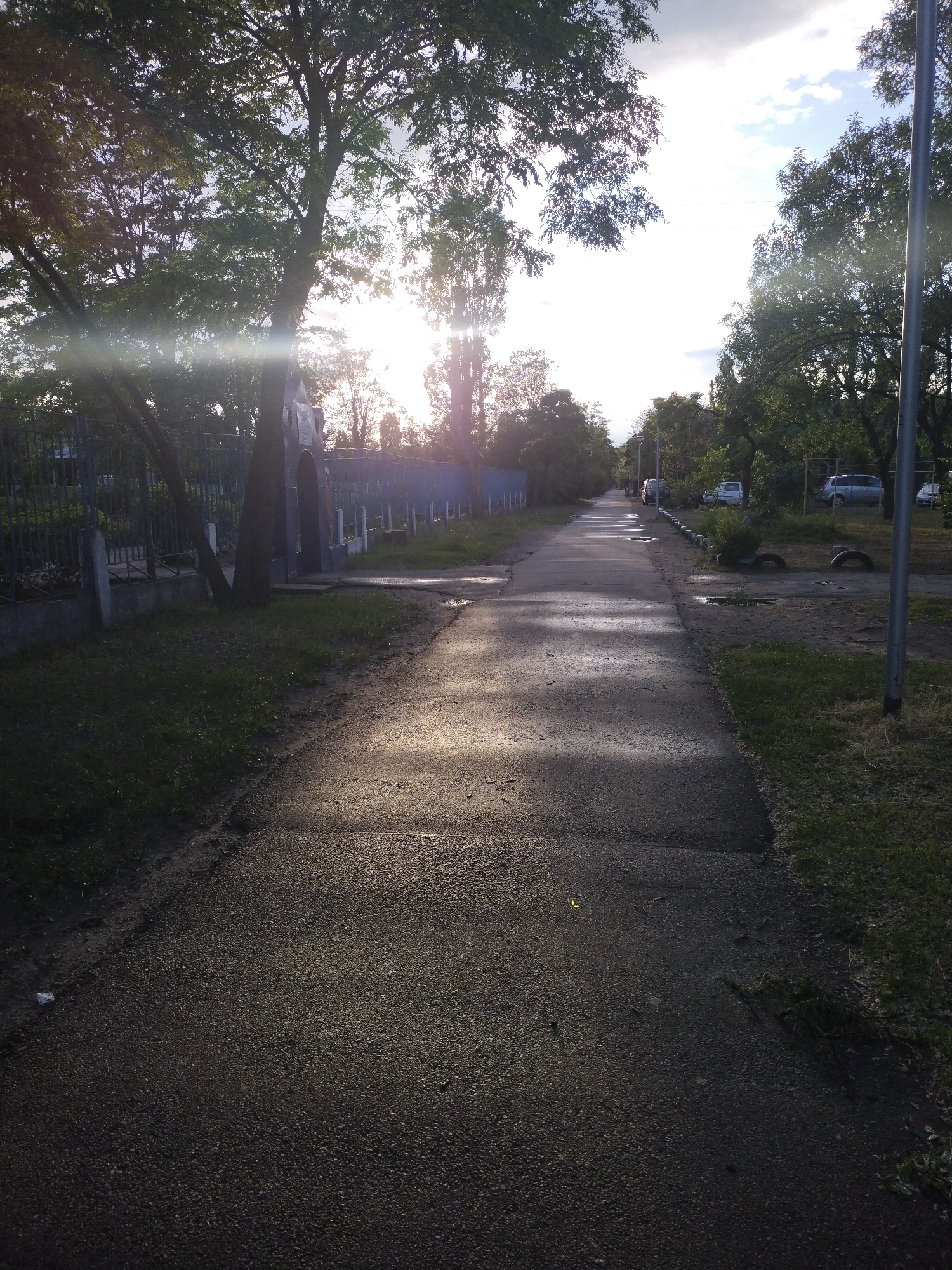 A ground-level, outdoor shot depicts a paved walkway receding into the distance, flanked by trees and a fence on the left, and trees and grass on the right. The walkway is dark, with areas of sunlight interspersed. The left side features a fence with metal posts and railings. Beyond the fence, more trees are visible. A dark-colored tree trunk is adjacent to the fence. The right side has patches of grass and more trees. In the distance, there are parked cars and other objects along the walkway. The lighting suggests either early morning or late afternoon. The sun is visible through the trees, creating a lens flare effect. The specific location is Horishni Plavni, Ukraine.