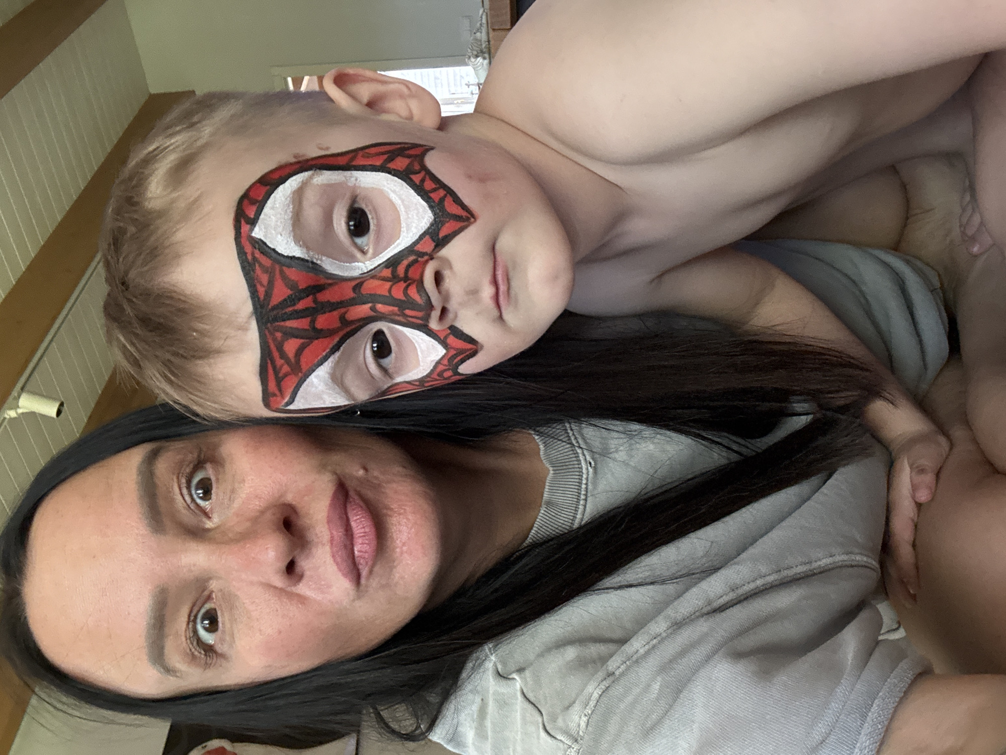 A selfie-style image shows a woman and a young boy lying down, likely indoors. The woman has dark hair and is wearing a light gray shirt. The boy is shirtless and has a Spider-Man mask painted on his face. They are close together, suggesting affection. The location is unknown; there is no information in the image to determine it is in Chaikovsky, Russia. The background is somewhat blurry but shows what appears to be a wall and some light fixtures.