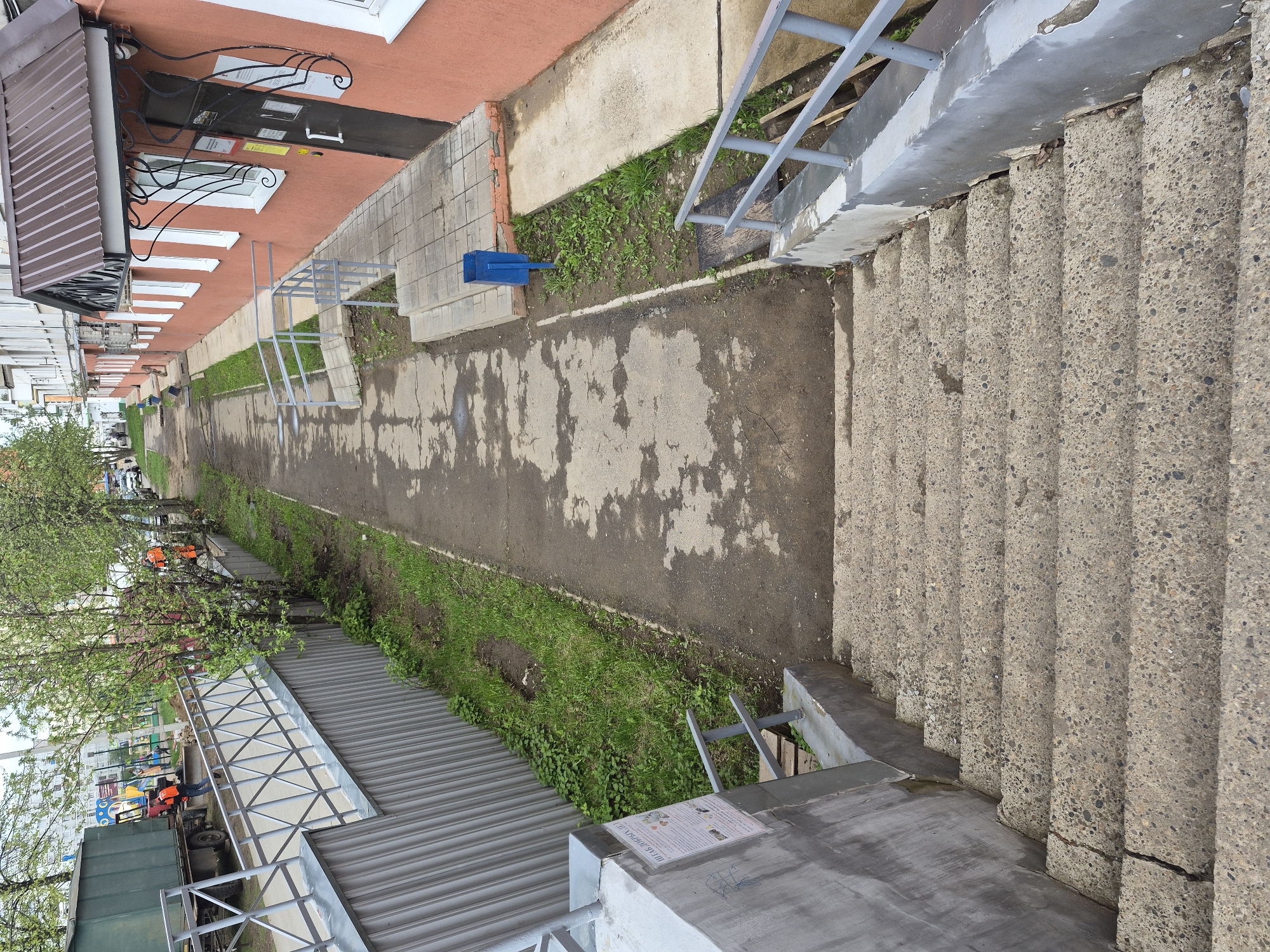 The image shows a view from above, likely from a building, looking down at an outdoor area. The right side features a set of concrete stairs with metal railings. Adjacent to the stairs is a walkway with patchy concrete and dirt areas, some with green vegetation growing. On the left side, there is a building with a pinkish facade and white-framed windows. Further left, there are trees with green leaves partially obscuring a view of other buildings and possibly vehicles in the distance. Closer to the viewer on the left, there's a corrugated metal structure with scaffolding-like metal frames and what appears to be a paper sign attached. There are no visible people in clear focus. The overall impression is of an urban, possibly industrial or mixed-use area. The location is in the city of Unknown, Unknown.