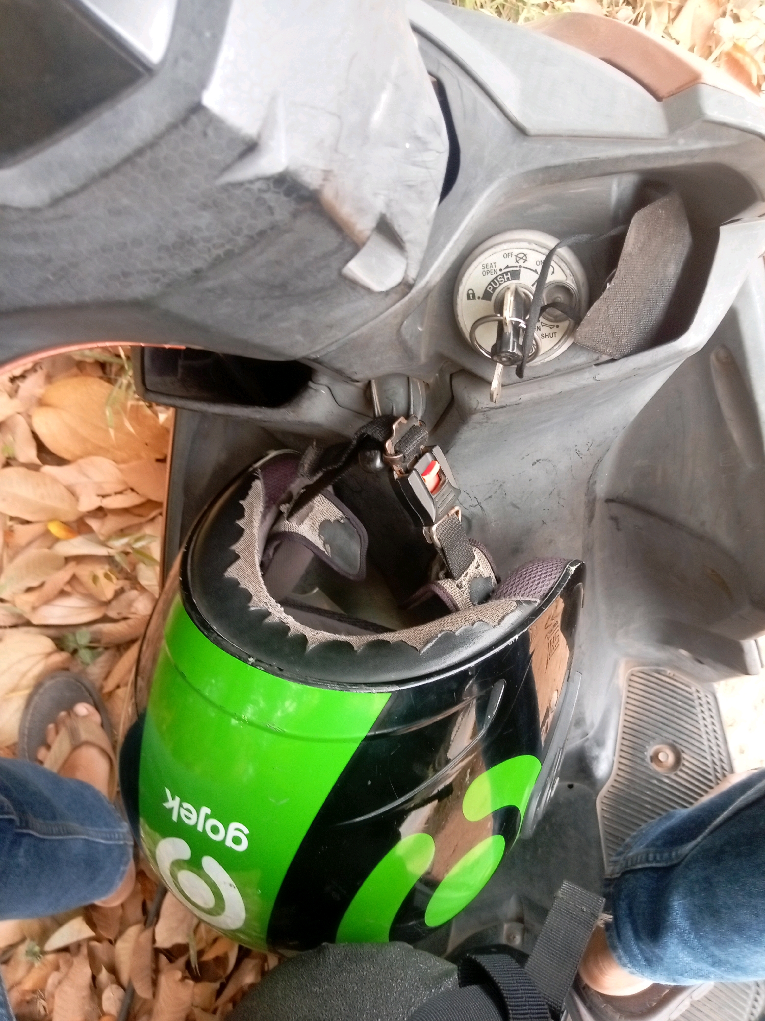 The image captures a close-up view of the front of a scooter, likely parked outdoors in a sandy or earthy area with scattered dry leaves, suggesting a location in Batam, Indonesia, possibly a roadside or a less maintained parking spot. A green and black Gojek helmet is positioned in the storage compartment of the scooter, with its strap secured. The scooter's ignition key is in the lock, set to the "OFF" position, indicating the vehicle is not currently in use. The rider's left leg and foot are visible on the left side of the frame, wearing blue jeans and a sandal, while the rider's right leg and foot are seen on the right side, wearing blue jeans and partially obscured by the scooter's floorboard. The overall scene suggests a moment of pause or rest, with the scooter parked and the rider not actively engaged with it.