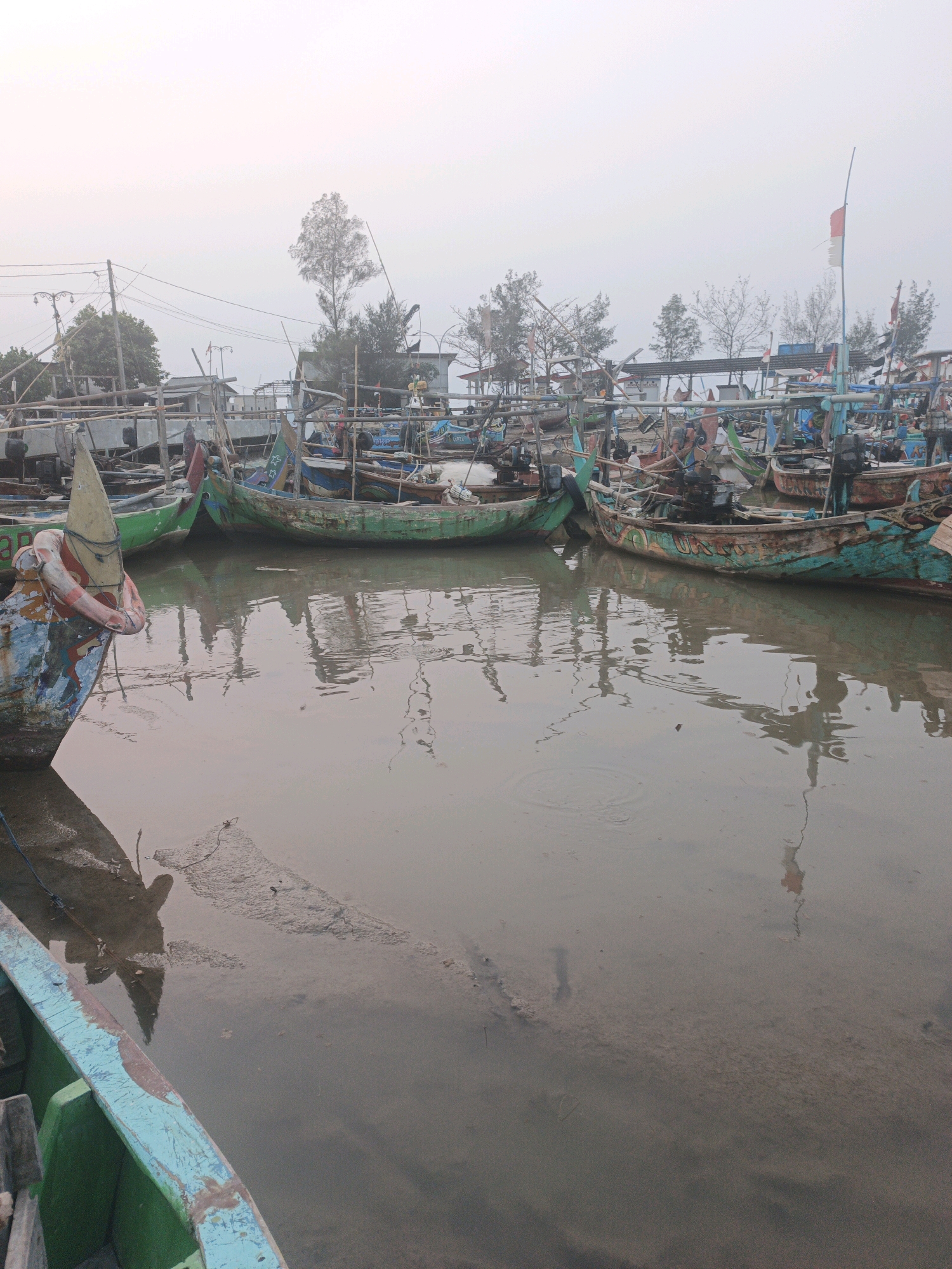 An aerial view shows a fleet of fishing boats docked in a shallow, muddy harbor. The boats are varied in size and color, with many painted in shades of green, blue, and red. Some boats are tied to wooden structures, while others are simply anchored. The water is murky and reflects the boats and the sky, creating a hazy, dreamlike effect. In the background, a few trees and buildings are visible, suggesting a coastal village. The sky is overcast, with a soft, diffused light that casts a muted glow over the scene. The overall impression is one of quiet stillness and maritime activity.
