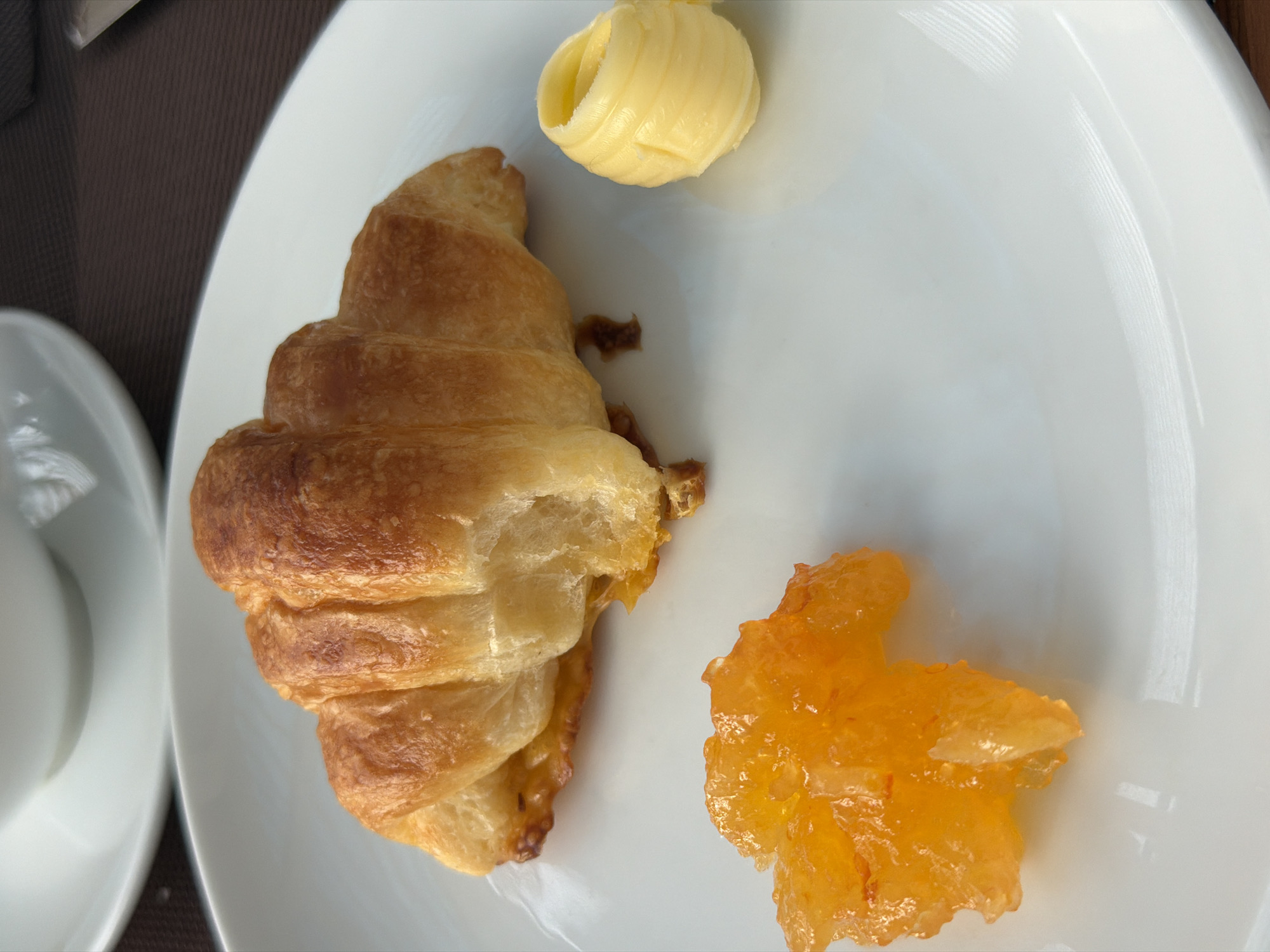 A close-up shot shows a white plate with a croissant, orange marmalade, and a small roll of butter. The croissant is golden brown and flaky, with layers visible. The marmalade is bright orange and translucent, with visible pieces of fruit peel. The butter is a pale yellow and appears to be a rolled curl. The plate is clean and white, and the background is a blurred brown surface, possibly a tablecloth. The lighting is bright, casting soft shadows. The scene appears to be a breakfast setting, and the image is taken from a slightly elevated angle.