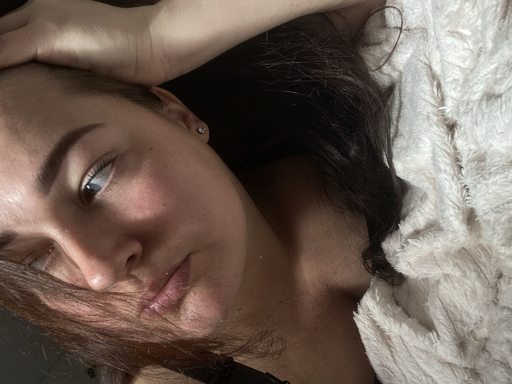 A woman is lying on a white, furry blanket. She has dark hair and is wearing a dark top. Her head is tilted to the left, and her left hand is resting on her forehead. The background is dark. There is no evidence suggesting the location of the image being in Sochi, Russia.