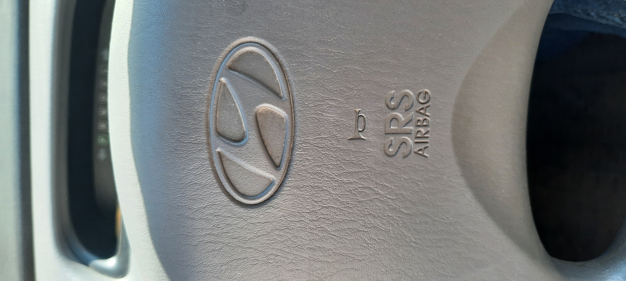 A close-up, eye-level, vertical shot focuses on the center of a car's steering wheel, specifically the airbag cover. The steering wheel appears to be made of grey, textured plastic. Embossed in the center is the oval-shaped Hyundai logo, with its characteristic stylized 'H'. Below the logo, also embossed, are the words "SRS AIRBAG" stacked, with "SRS" above "AIRBAG". A small, stylized horn symbol is visible above the "SRS" text. The rest of the steering wheel and the surrounding dashboard are out of focus and not clearly discernible. The lighting suggests an interior shot, possibly during daytime due to the brightness and subtle shadows. There are no people visible in the image, and no context is provided to place it in Astrakhan, Russia.