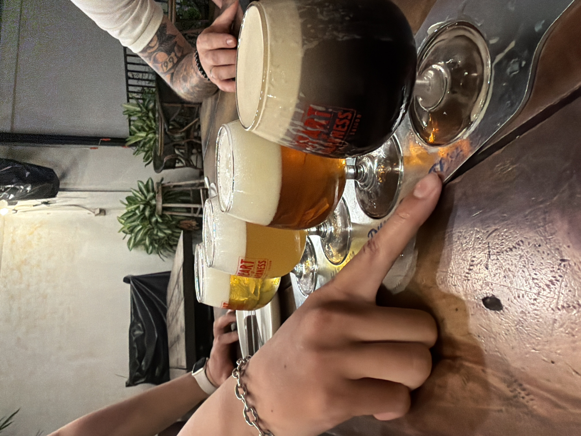 The image shows a person pointing at a flight of four beers on a metal serving tray, set on a wooden table. The beers vary in color from light yellow to dark brown/black. A second person with tattooed arms is partially visible in the top left corner. The location is Nha Trang Ward, Vietnam, as indicated by the beer brand "Heart of Darkness" which has a location in this city. The background shows plants, a black bag, and a white wall.