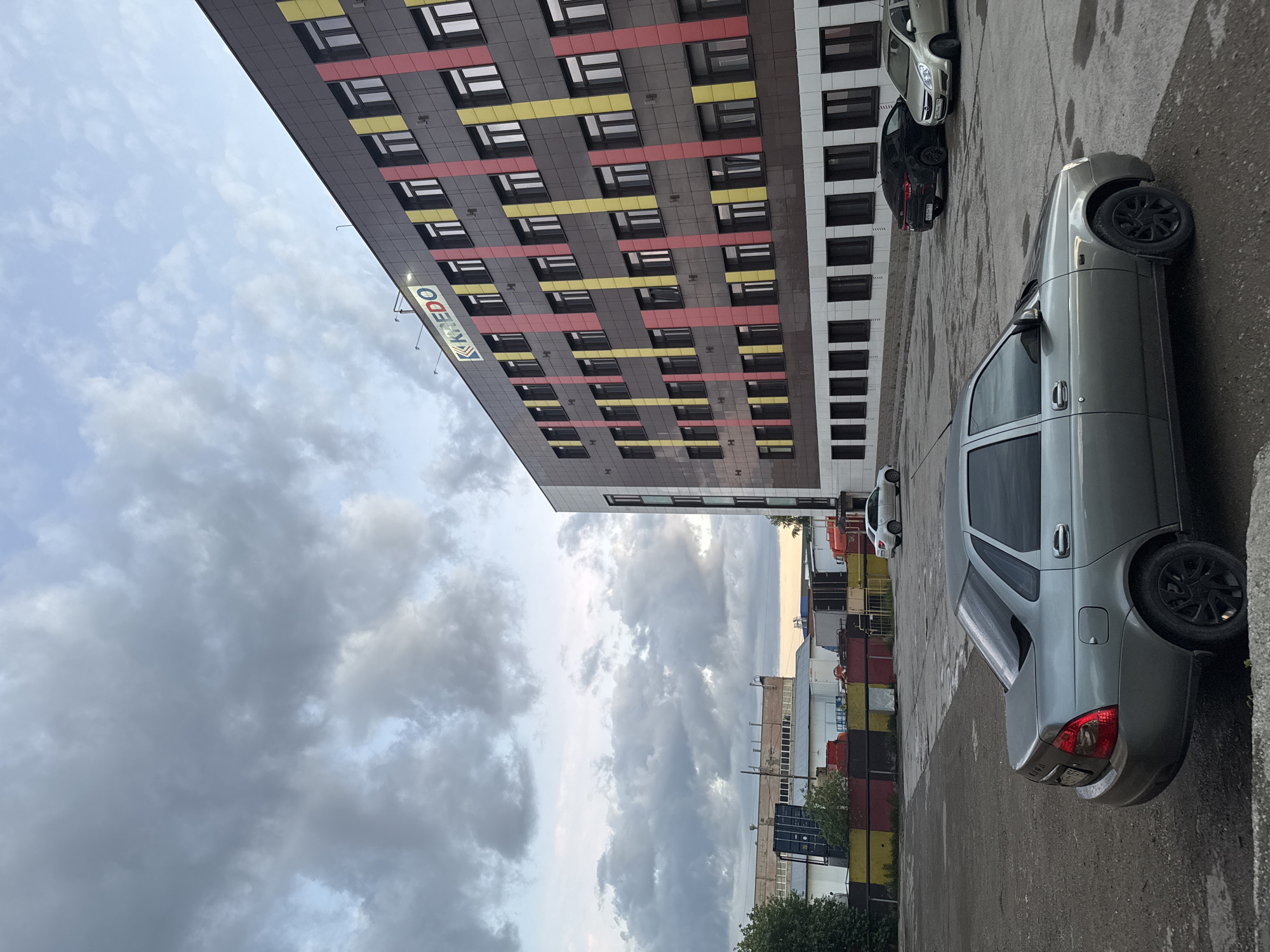 The image captures an urban street scene in Perm, Russia, featuring a modern building, several parked cars, and an overcast sky. The primary subject is a tall, contemporary building on the left, characterized by its dark grey facade. This facade is visually distinctive with horizontal bands of yellow and red color alternating with numerous rectangular windows. A sign with the blue text "KREDO" is prominently displayed on the side of this building.
In the foreground and towards the right, a silver sedan is parked on an asphalt lot. The rear of the car shows the "LADA" emblem, and it is fitted with black multi-spoke wheels. Further back, a dark-colored car and another light-colored vehicle are also parked.
The sky above is heavily overcast with grey clouds, indicating a cloudy day, possibly late afternoon or early evening given the ambient light. In the distance, beyond the main building and cars, other commercial or industrial structures are partially visible under the cloudy sky. There are no people visible, and no specific activities are taking place in this static depiction of a city environment.