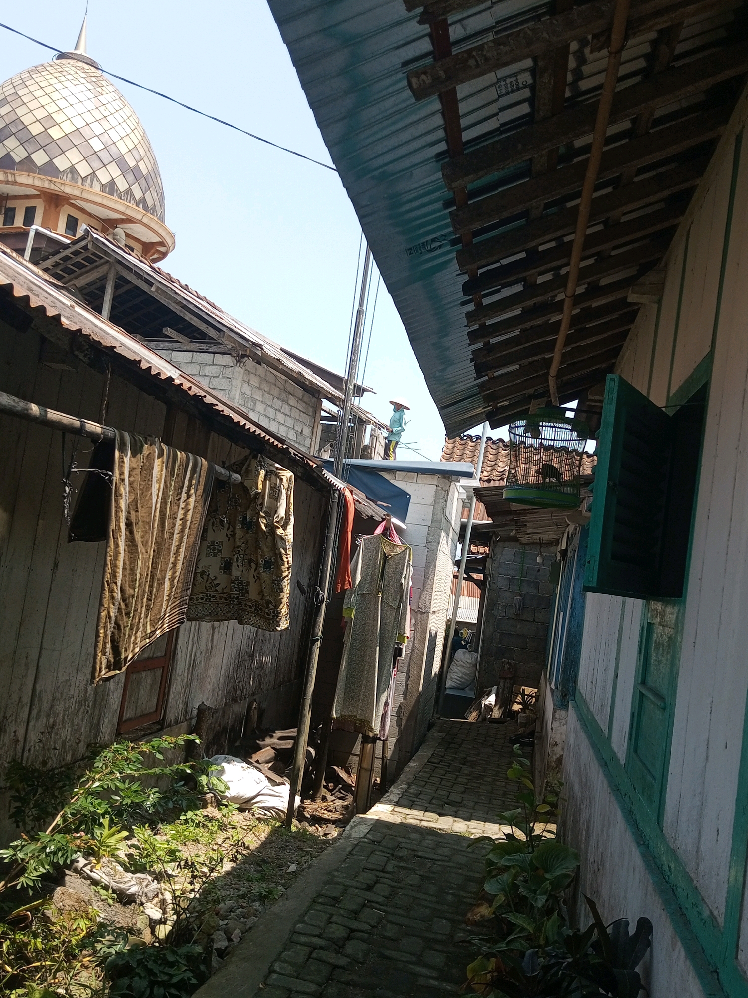 The image captures a narrow, sun-drenched alleyway in Andongsili, Indonesia. A mosque with a distinctive dome is visible in the upper left background, partially obscured by other structures. Buildings with weathered wooden siding and corrugated metal roofs line the alley. Laundry hangs on a clothesline on the left side of the alley, featuring patterned fabrics. On the right, a white building with green trim features an open window. A green birdcage hangs from an overhead structure, containing two birds. A person wearing a conical hat stands on a rooftop in the distance. The alley itself is paved with cobblestones, and vegetation grows along the sides. Sunlight casts strong shadows, indicating a bright, sunny day. The overall impression is one of a rural or semi-rural residential area with traditional architecture.