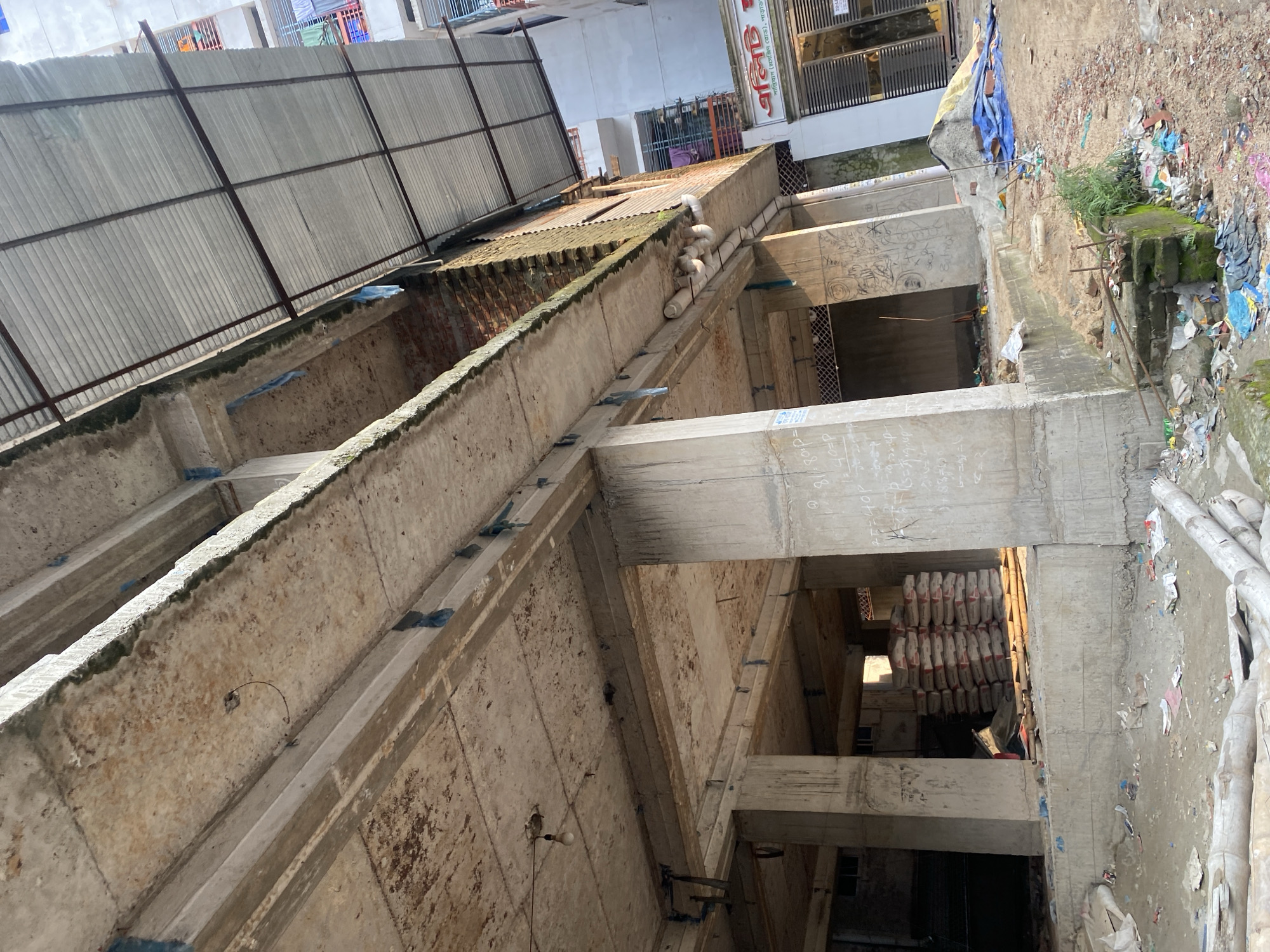 The image shows the construction site of a building, likely in Dhaka, Bangladesh. The exposed concrete framework reveals multiple levels, with scaffolding and construction materials, including bags of cement, visible within the structure. The site appears unfinished, with debris and trash scattered on the ground. The surrounding area includes other buildings, one of which displays signage with Bengali script.