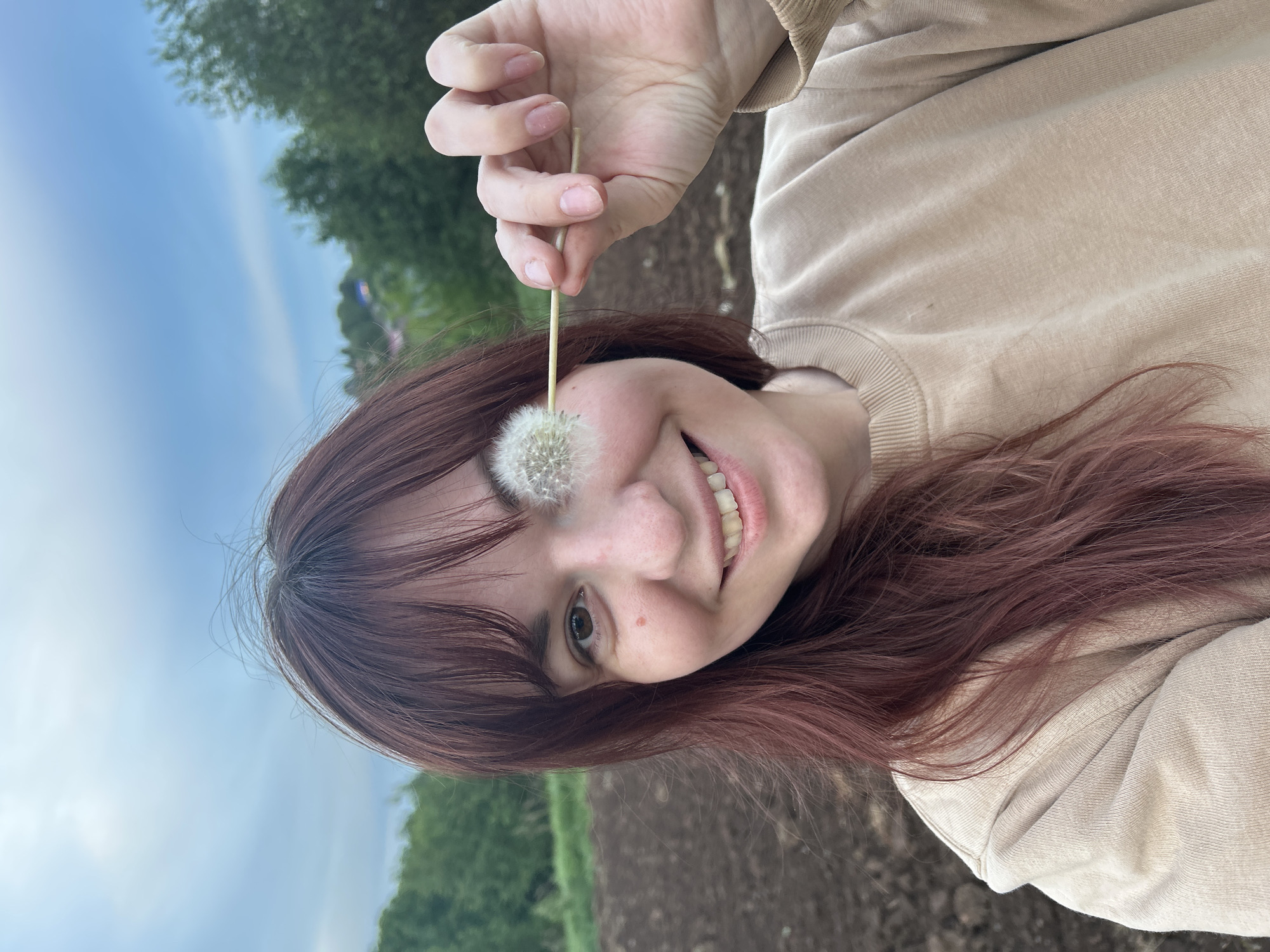 A reddish-haired woman is holding a dandelion in front of her right eye while smiling, facing the camera. She is wearing a beige crew neck sweatshirt. She stands in a field near Фоки, Russia. Behind her are trees, open sky, and a cultivated field.