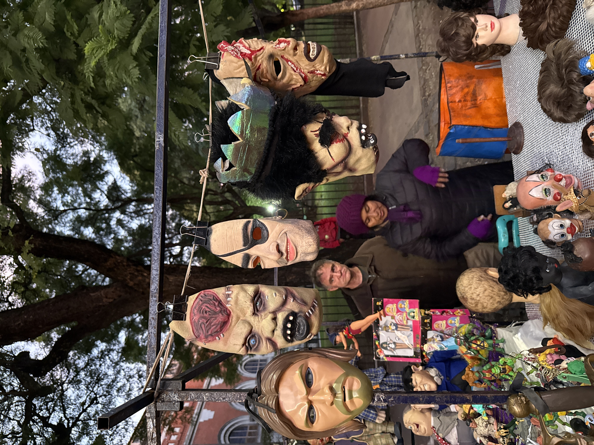 The scene depicts a market stall in a public space, likely in Buenos Aires, Argentina given the architecture visible in the background. The stall is displaying a variety of items, primarily Halloween-style masks hanging from a metal frame. The masks are grotesque and depict various monsters and disfigured faces. Below the masks, there is a collection of dolls, figurines, and other toys. Two adults, one female wearing a purple hat and a grey jacket and one male, are partially visible behind the stall, seemingly attending to it. The lighting suggests it is late afternoon or early evening. Trees are also visible in the background.
