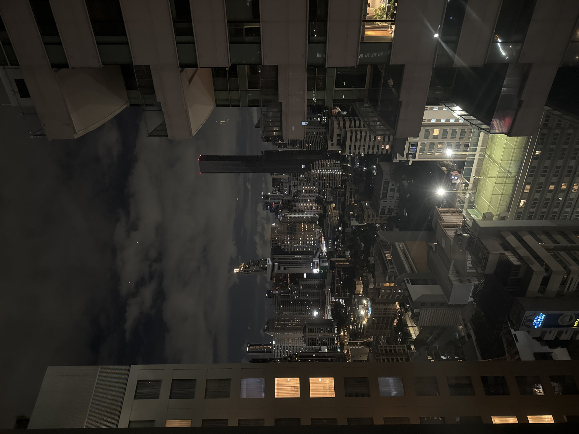 The image depicts a nighttime cityscape, seemingly photographed from an elevated position, likely a hotel room, offering a view of Bangkok, Thailand. The photograph is oriented upside down. Various high-rise buildings dominate the scene, illuminated by artificial lights. Skyscrapers of varying heights and architectural styles are visible throughout the city. A tall, prominent building with lighted top is centrally positioned. The dark sky is scattered with clouds. A rooftop tennis court is visible. The upper and lower edges of the image are framed by what appears to be the window frame or the architecture of the building from which the photograph was taken. A crescent moon is visible in the sky. The overall atmosphere is urban and dark, with artificial light illuminating the buildings.