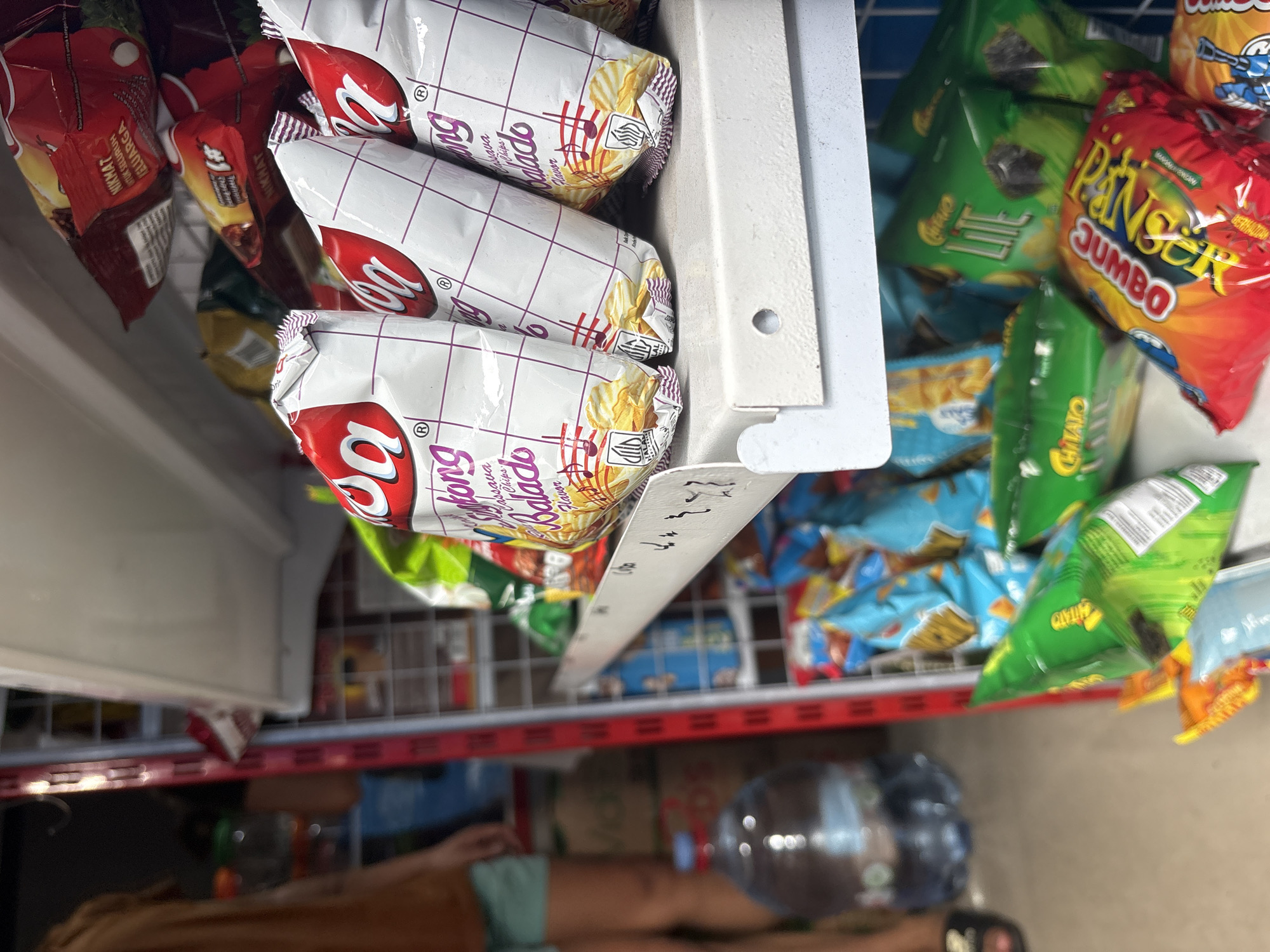 The image shows a close-up view of shelves in a convenience store, likely located in North Jakarta, Indonesia. The shelves are stocked with various snack food items, predominantly bags of chips. In the foreground, several bags of "Kopi Luwak" brand chips, described as "Cassava Chips Balado Flavor," are prominently displayed. These bags are white with red and purple accents. To the right and below, other chip bags are visible, including green bags of "Chitato Lite" and a red bag with yellow text reading "Piknik Jumbo." In the background, a person, partially obscured, is seen standing near a large water dispenser. The overall environment suggests a typical retail setting with merchandise neatly arranged on shelves.
