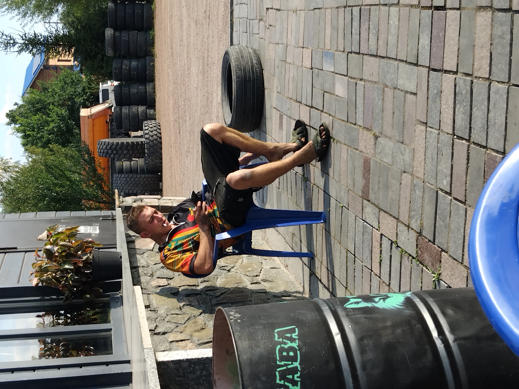 The image depicts a man sitting on a blue plastic chair outdoors, with a cigarette in his mouth, looking at his phone. He is wearing a colorful, patterned short-sleeved shirt and black shorts. His legs are bare and he is wearing dark sandals. In the foreground to the left, a large black barrel with green text and a green graphic of an animal is visible. Behind the man, a large black tire is lying on the ground. In the background, stacks of tires are arranged, a dump truck, and a building with a blue roof can be seen. The ground is paved with rectangular bricks. The scene appears to be in an outdoor industrial or storage area, possibly a tire shop or a junkyard. The presence of the building with a blue roof and the general landscape suggests a location in a city or town in Ukraine, potentially in the city of Novoselytsia.
