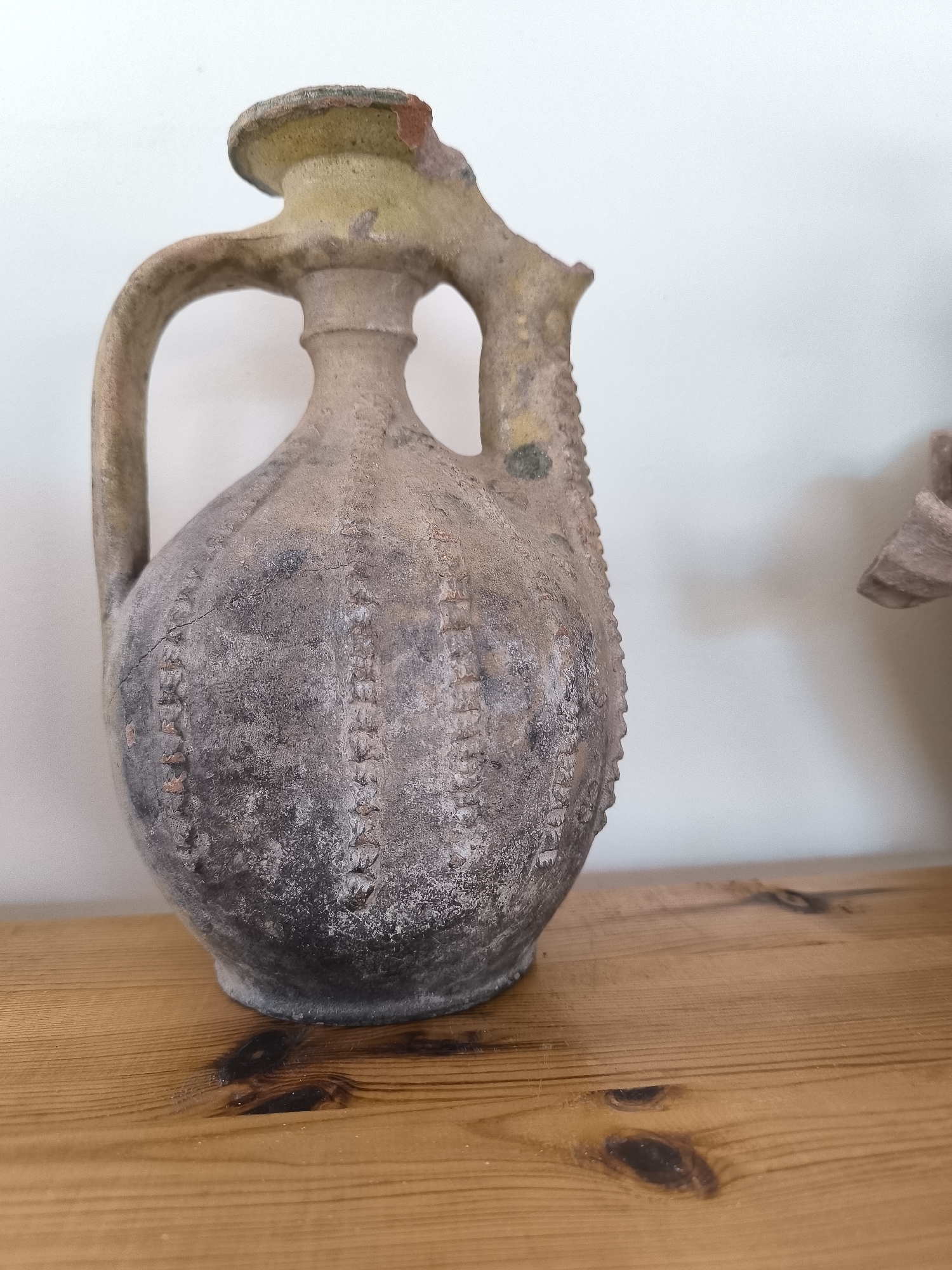 An ancient amphora with a distinctive ribbed texture is displayed on a wooden shelf. The amphora is primarily earthen-toned with areas of grey and black patina, suggesting age and possibly mineral deposits. It has two handles arching over its opening and a narrow neck leading to a bulbous body. The rim of the amphora appears to be chipped in places, revealing a lighter inner layer. The surface of the amphora is cracked in several areas, with some fragments missing. The wooden shelf is made of light-colored planks with visible knots. The background is a plain white wall. The amphora is located in Garmen, Bulgaria.