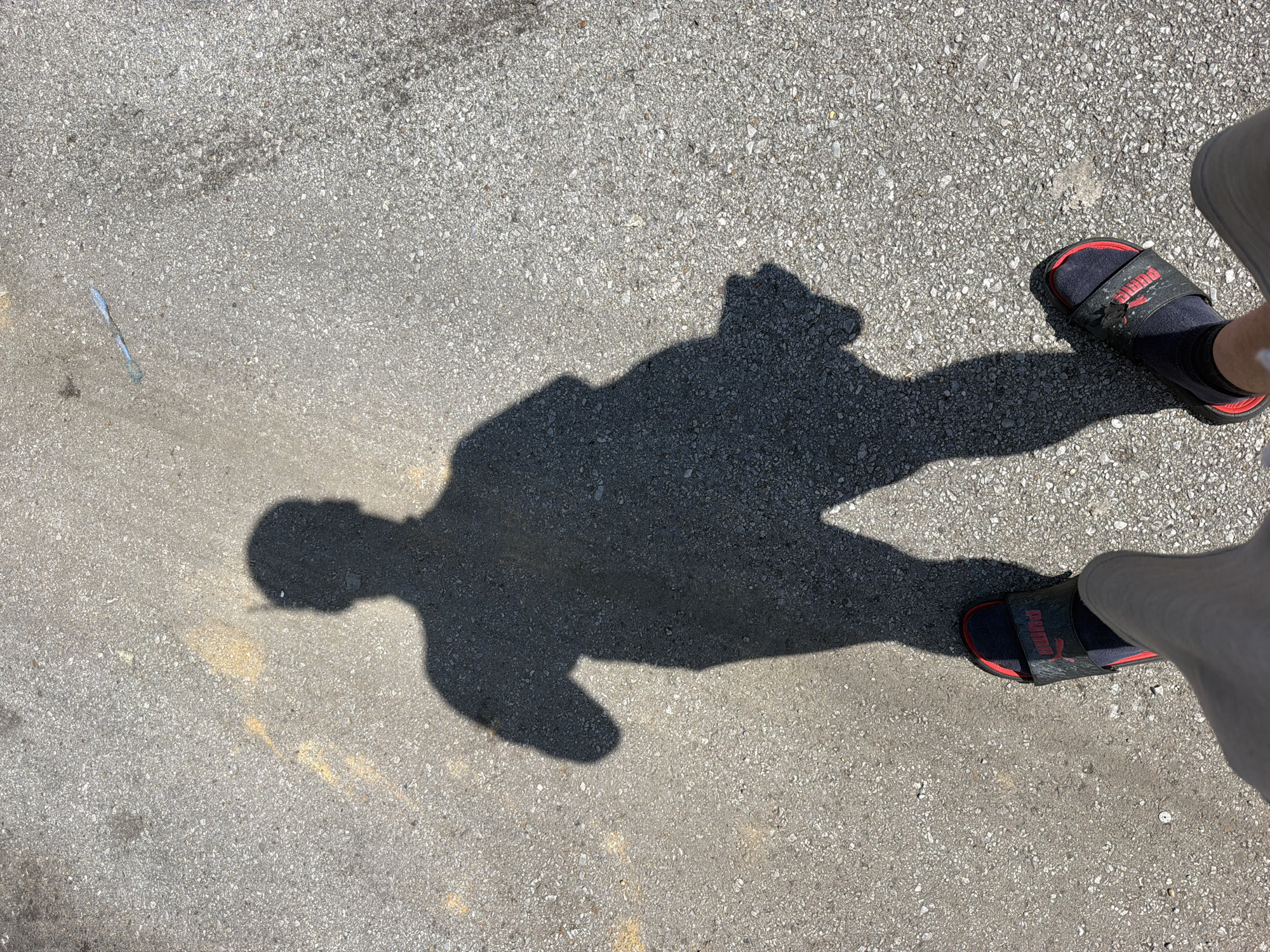 The image shows a shadow of a person standing on asphalt. The shadow is cast by the sun, indicating it is daytime. The person is wearing sandals. A thin, light blue object is visible on the asphalt to the right of the shadow. The ground appears to be a road or parking lot, with faint yellow lines visible. The location is the city of Lebanon, United States of America.