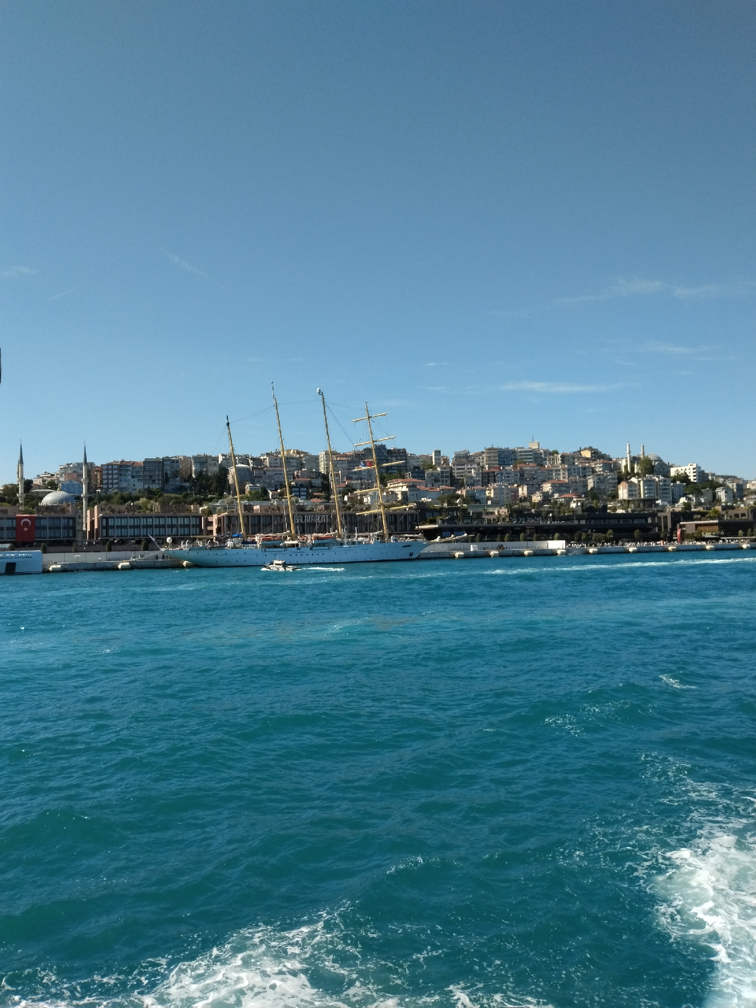The image shows a waterfront scene in Sarıyer, Turkey, featuring a large white sailing ship docked along the shore. Behind the ship, a cityscape is visible, characterized by numerous buildings of varying heights and architectural styles. A mosque with minarets is present on the left side of the image. The foreground is dominated by blue water, with visible wake patterns indicating movement. The sky is clear and blue.
