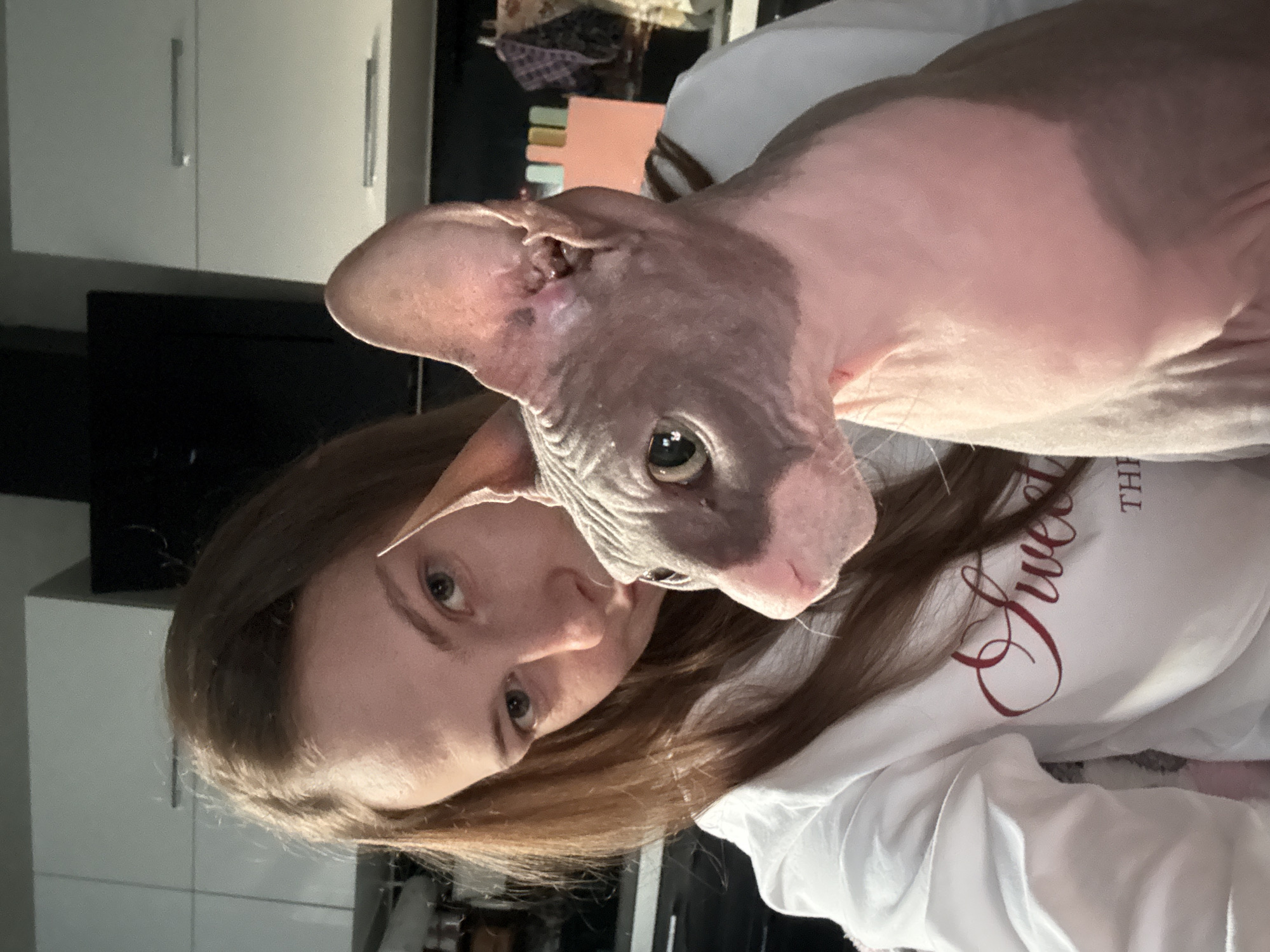 A selfie is taken by a young woman with long brown hair, wearing a white sweatshirt with red lettering. She is holding a hairless cat, a Sphynx breed, which has pink and gray coloring and large ears. The cat is looking to its right, while the woman looks at the camera. They are indoors, with kitchen cabinets visible in the background. The location is identified as Novaya Ivanovka, Russia.