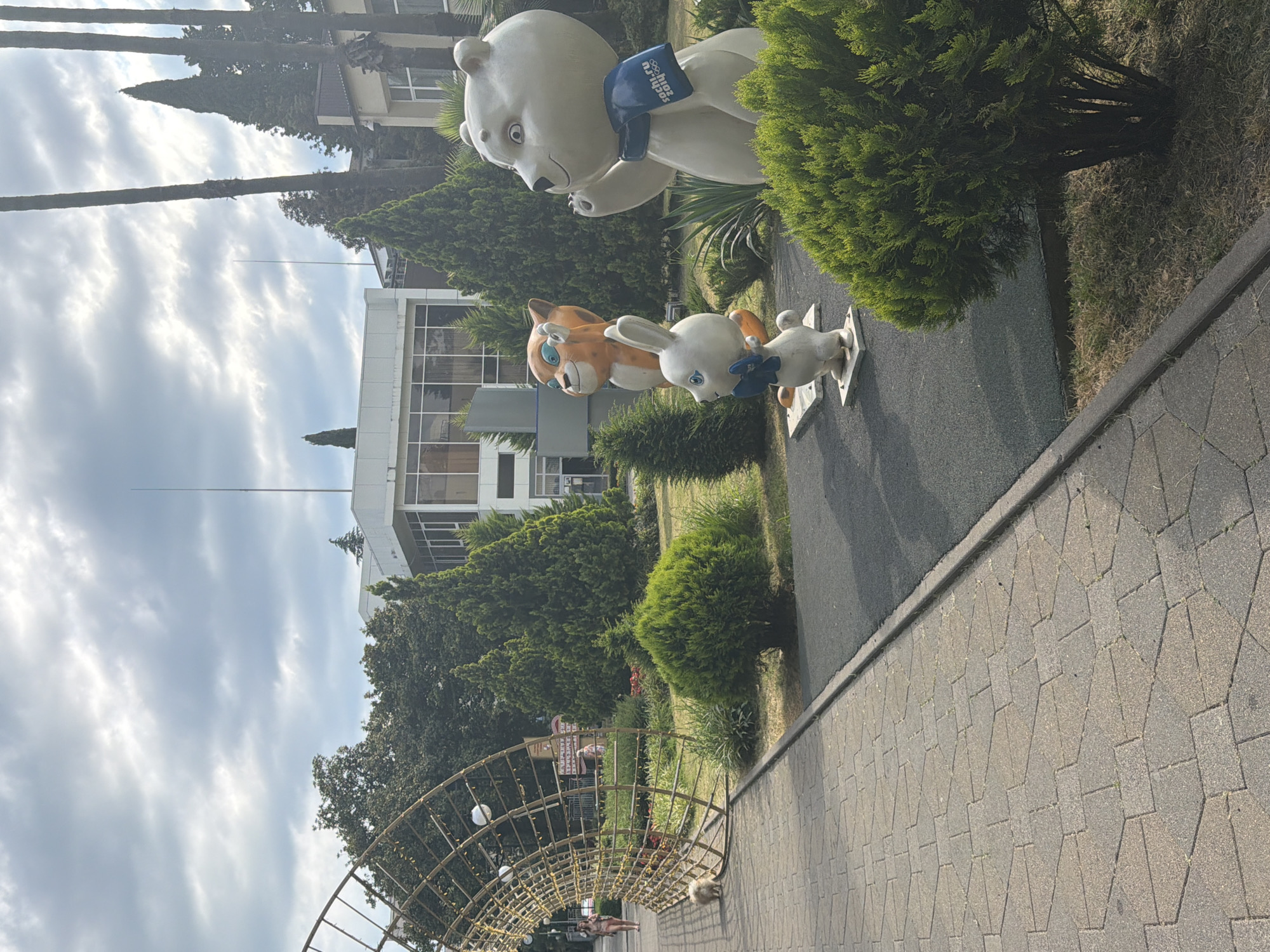 The image shows an outdoor scene in Sochi, Russia, featuring a paved walkway and a park area. In the park, there are three statues of cartoon characters: a white bear, a white rabbit, and an orange tiger. The bear statue is the largest and is wearing a blue scarf with "Sochi.ru 2014" written on it. The rabbit is in front of the tiger, and both are smaller than the bear. To the left of the statues, a small, fluffy dog is walking on the paved path. In the background, there is a large, arched metal structure with lights, and beyond that, a building with large windows. Several trees, including palm trees, are visible throughout the scene, and the sky is partly cloudy. The overall impression is of a public park area in a resort town.