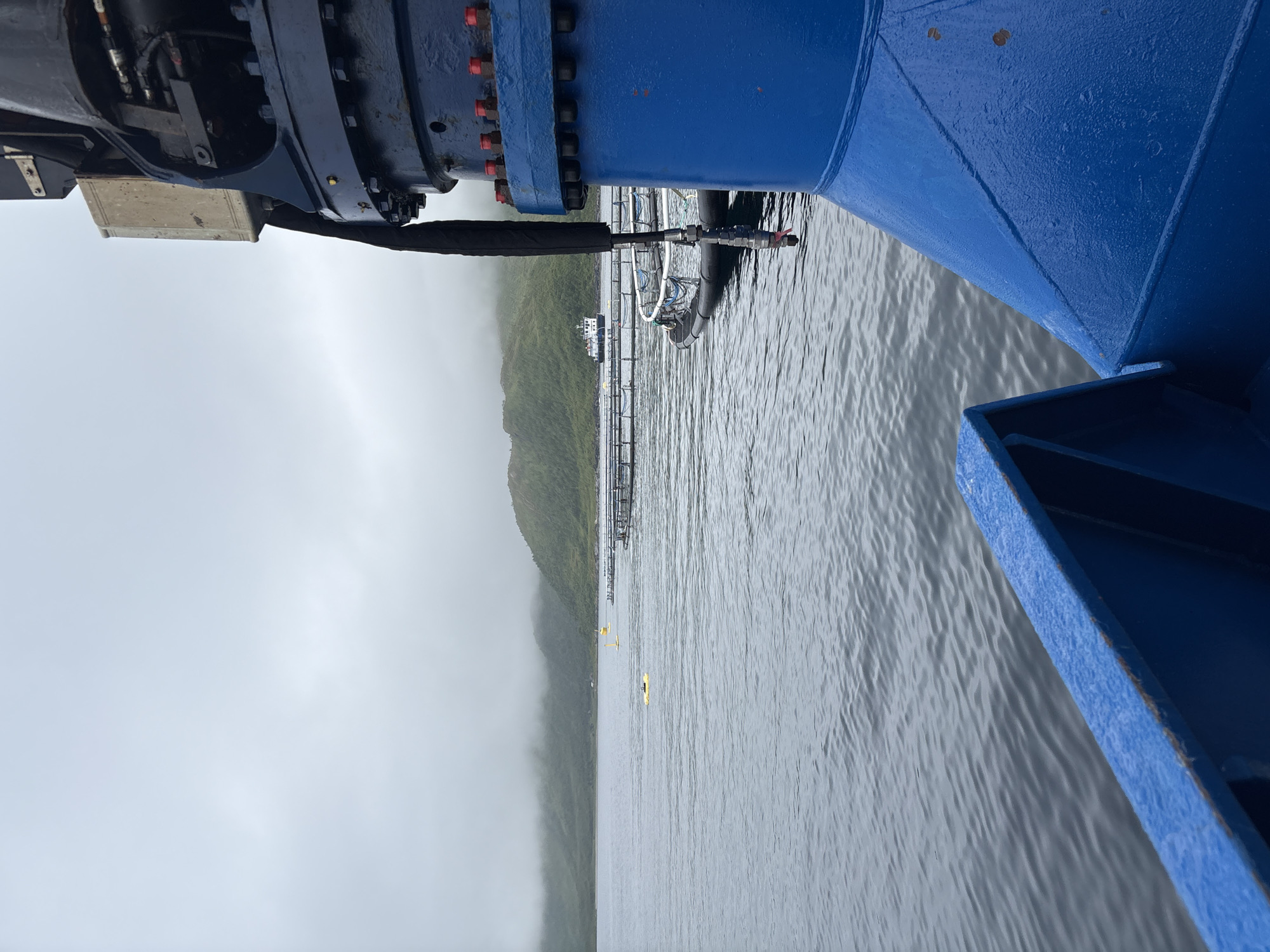 The image depicts a calm body of water with a misty, overcast sky. In the foreground, a large blue industrial object, likely part of a crane or machinery, dominates the right side of the frame. The water is a dark gray with small ripples. In the midground, a series of aquaculture pens are visible, marked by buoys and netting. A small boat is also present in the distance, near the pens. The background shows a sloping, green hillside shrouded in fog. The scene appears to be a section of the harbor or a fjord near Botnhamn, Norway, given the presence of aquaculture and the typical coastal landscape. No people are visible in the image.