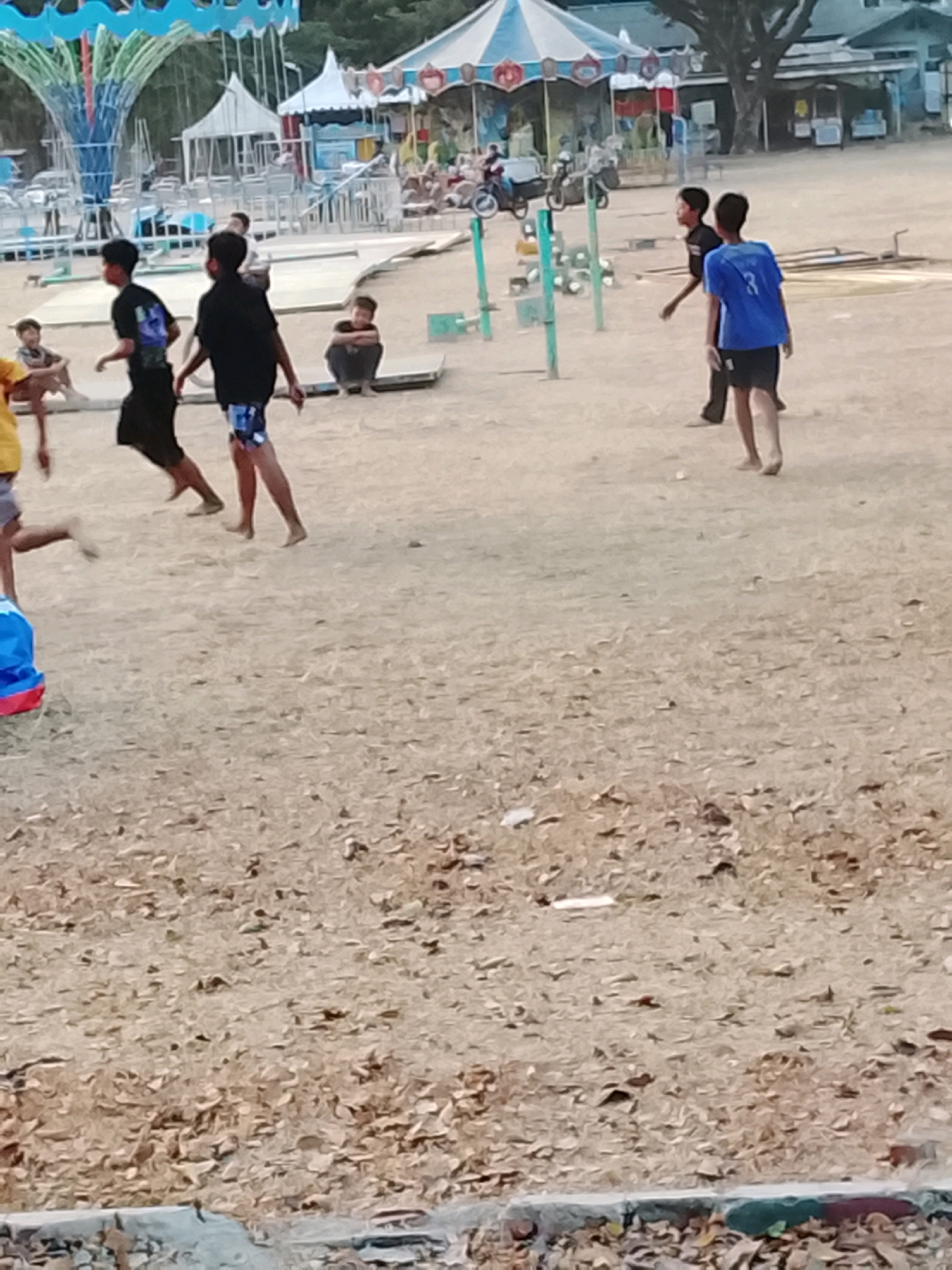 The image depicts a sandy outdoor area with a carousel in the background, suggesting a fairground or park setting. Several children and teenagers are present, engaged in various activities. In the foreground, two boys are running. One is wearing a black t-shirt and patterned shorts, while the other is in a black t-shirt and black shorts. To their right, a boy in a blue t-shirt with the number 3 on the back and black shorts is walking away from the camera. Further back, another boy in a black t-shirt and patterned shorts is sitting. A person in a yellow t-shirt is partially visible on the left. The environment is characterized by dry, sandy ground with scattered leaves. The overall scene suggests a casual, recreational gathering of young people in an outdoor public space. The location is identified as Harjosari Lor, Indonesia.