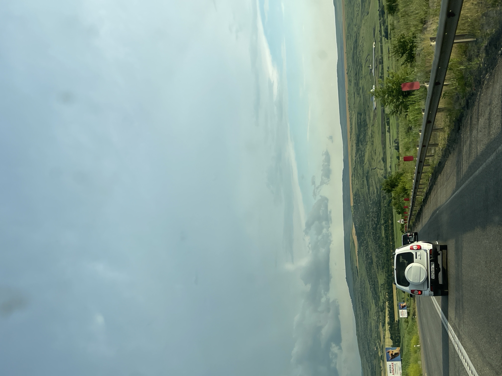This is a wide, slightly upward-angled, eye-level, outdoor, natural light, full shot photograph taken from a vehicle traveling on a paved road. The sky is cloudy, with a light blue hue and darker gray clouds on the left. Below the sky, rolling green hills and fields stretch into the distance, with a few farm buildings visible. The road has a white line on the left side and a metal guardrail with red markers on the right. In the foreground, a white SUV with a spare tire mounted on the back is driving on the road. The license plate on the SUV reads "K428KB 126". Two billboards are visible on the left side of the road. One is a large billboard with an image of a dog and text, and the other is a smaller billboard with a similar image and text. The scene appears to be in the countryside, likely in or near the city of Tatarka, Russia, given the Cyrillic license plate and billboard text.